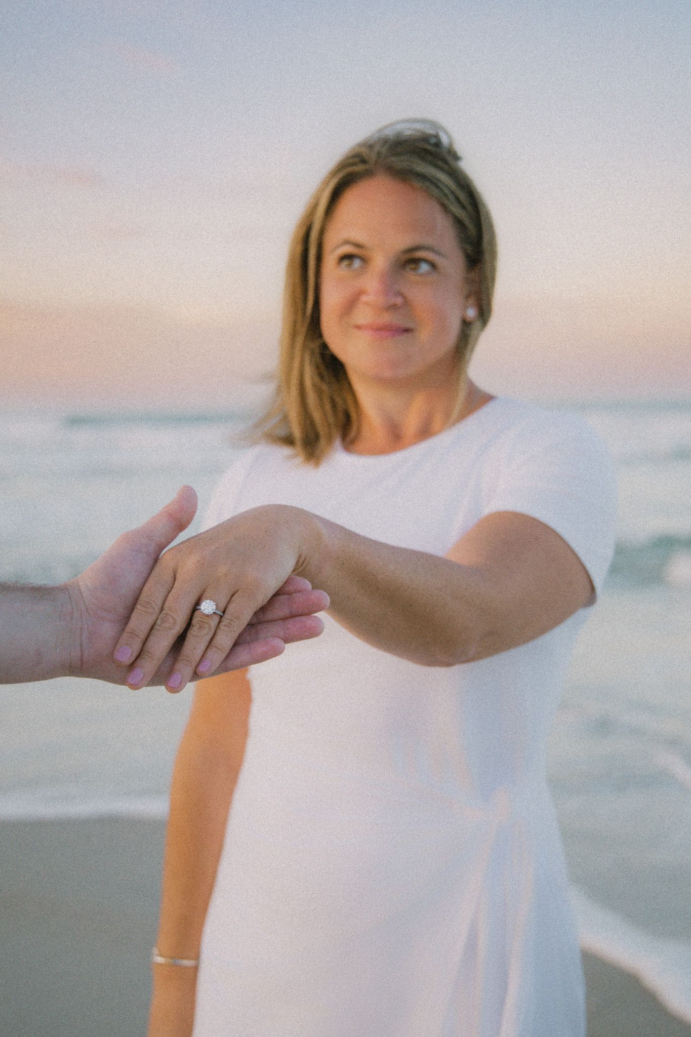 Woman in white dress on beach holding hands showing engagement ring at sunset.