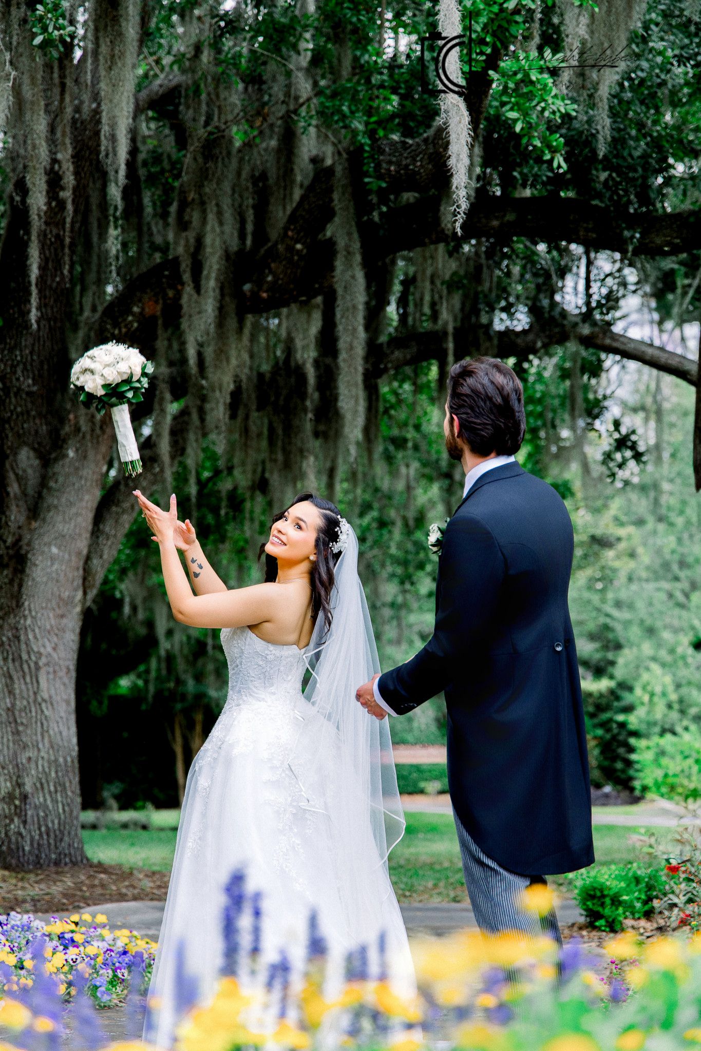 Bride tossing bouquet under Spanish moss trees while groom watches in lush garden wedding setting.