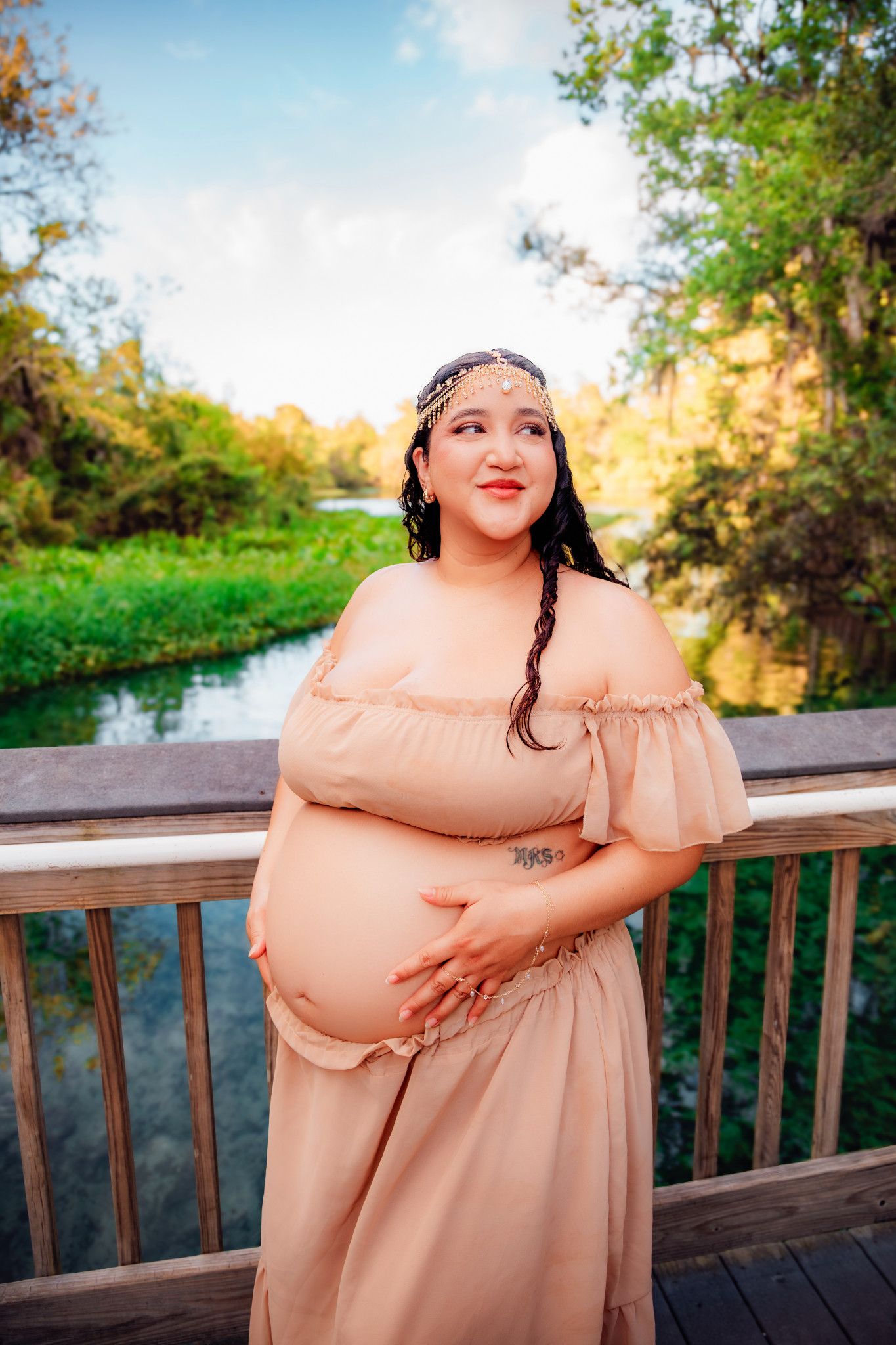 Pregnant woman in blush off-shoulder dress cradling baby bump on wooden bridge surrounded by lush greenery.