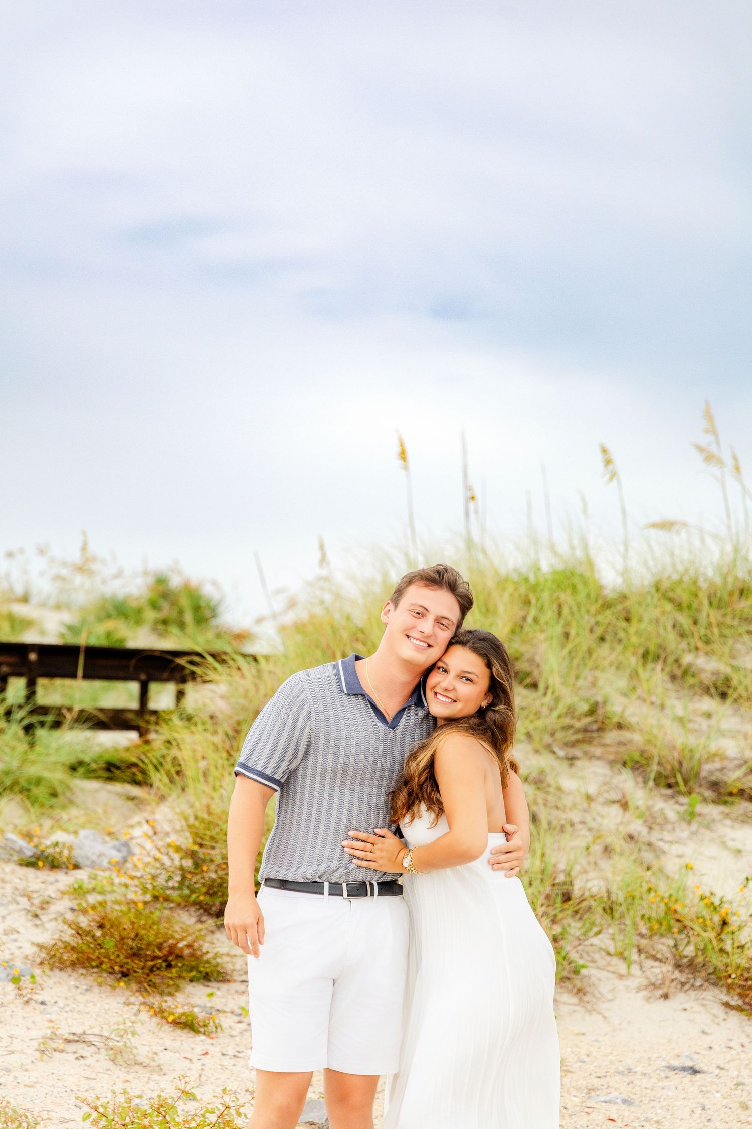 Happy couple embracing on sandy beach dunes, woman in white dress and man in striped shirt, smiling outdoors.