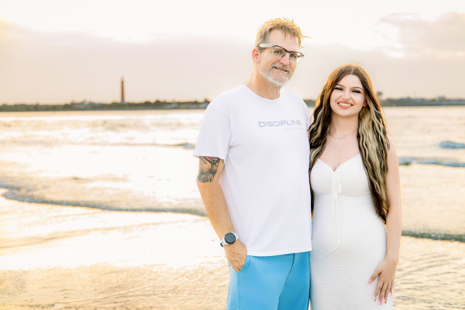 Couple posing together on a beach at golden hour with a lighthouse in the background.