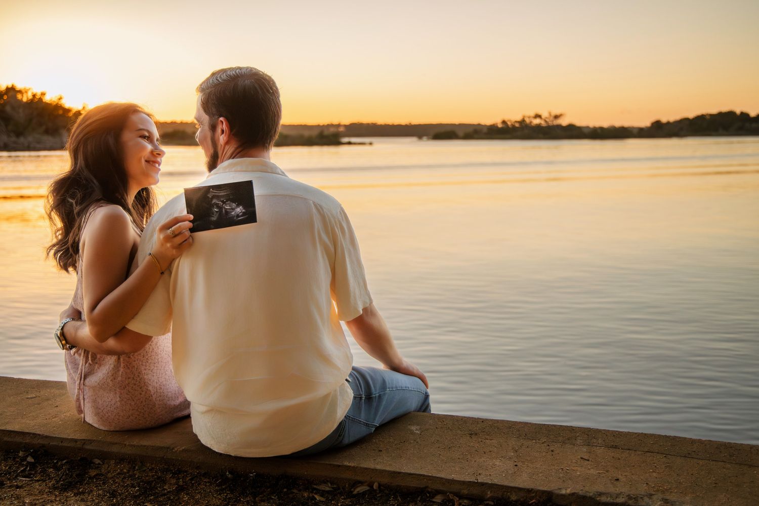 Couple sitting by a lake at sunset holding an ultrasound photo, announcing pregnancy in a romantic outdoor setting.