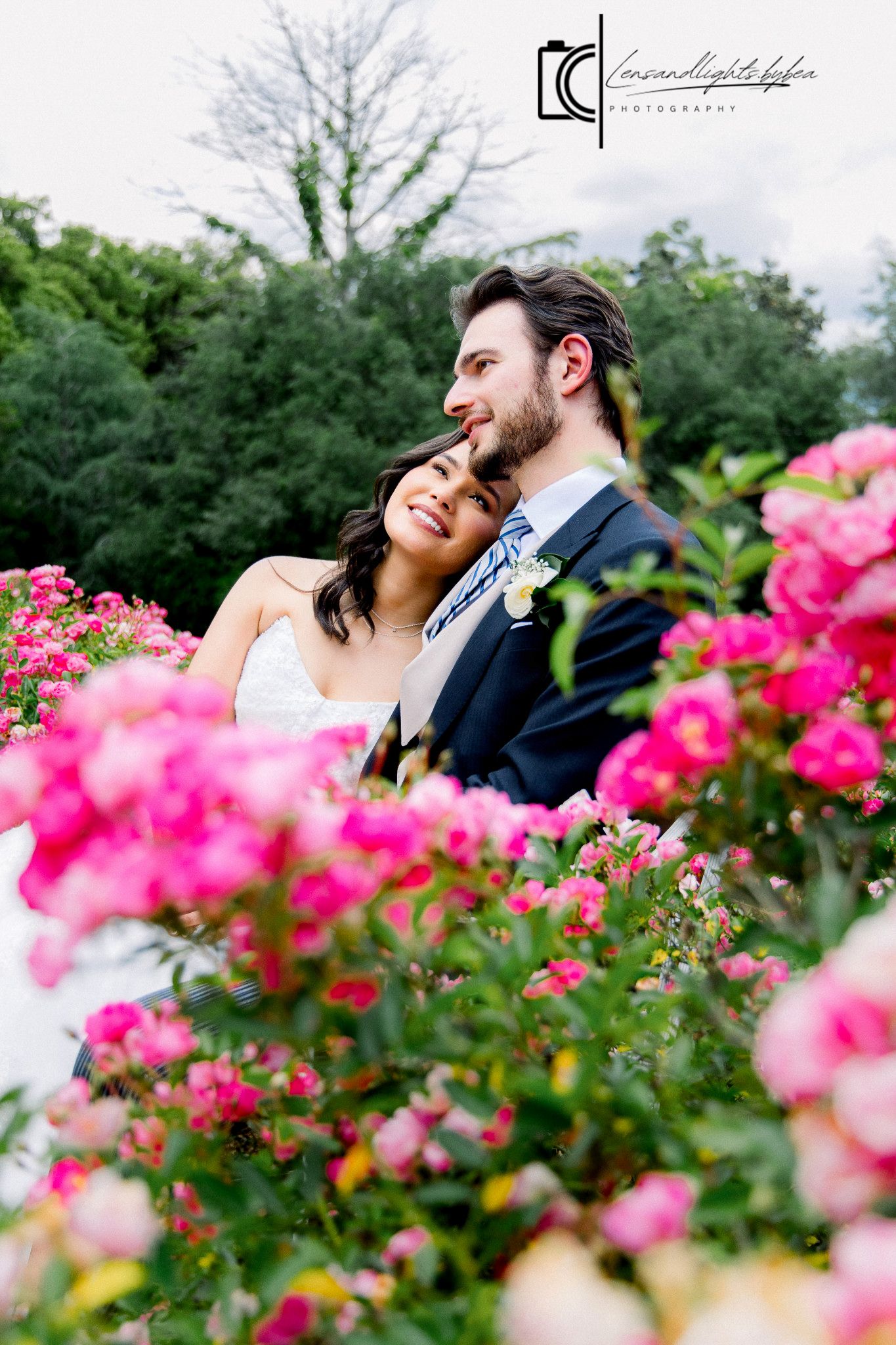 Wedding couple embraces among vibrant pink roses in a lush garden, groom in blue suit with boutonniere.