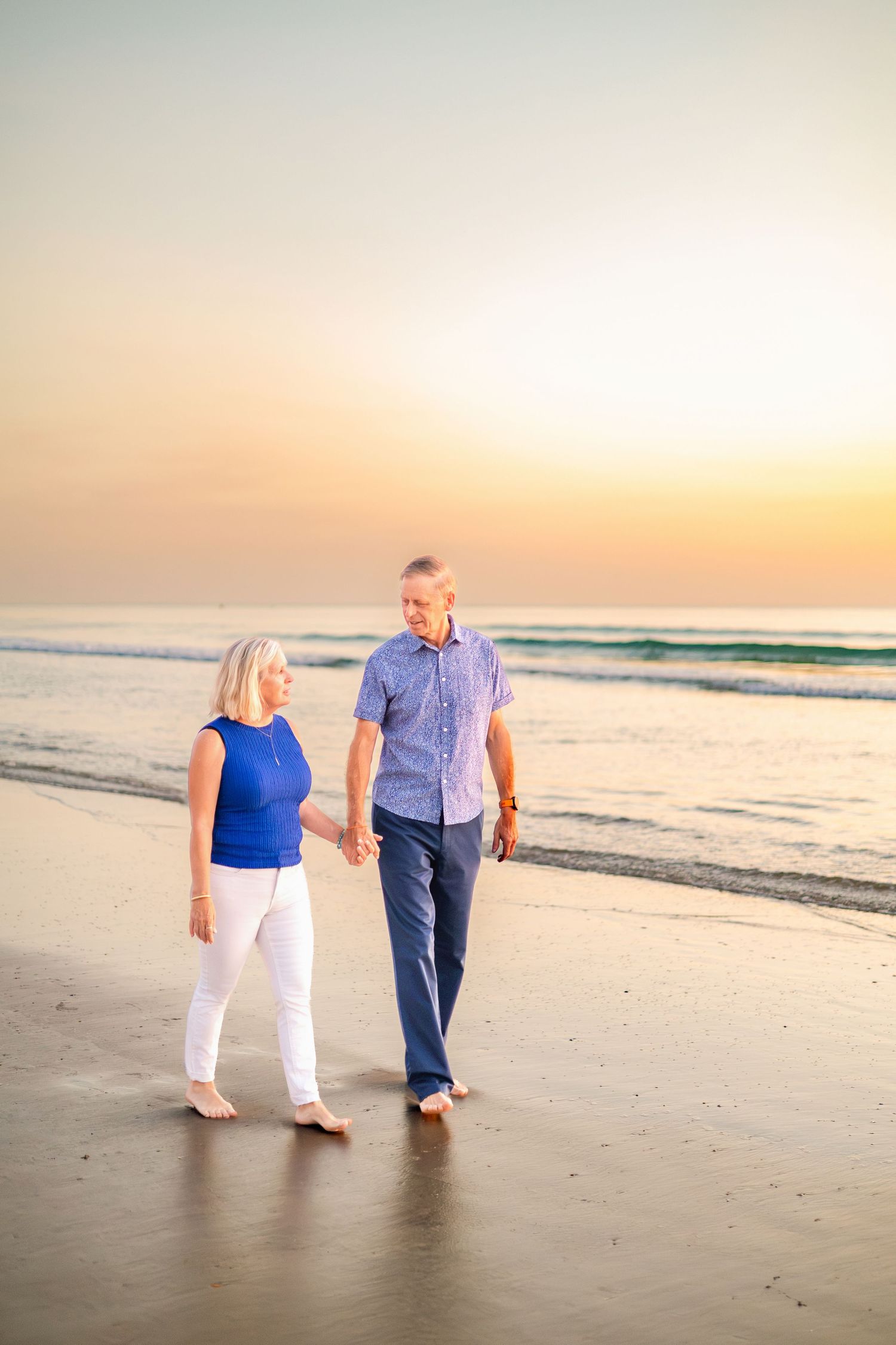 Mature couple holding hands walking barefoot on a sandy beach at sunset with ocean waves in the background.