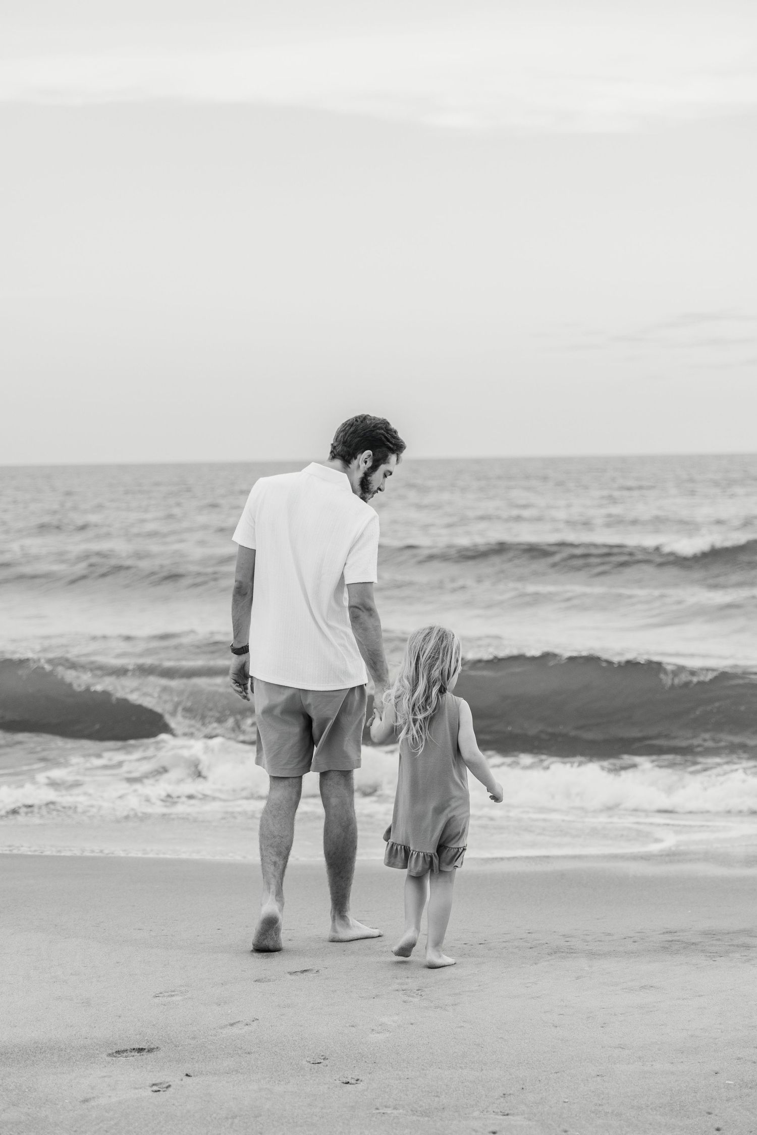 Black and white photo of a father and young child holding hands while walking barefoot on a sandy beach near ocean waves.
