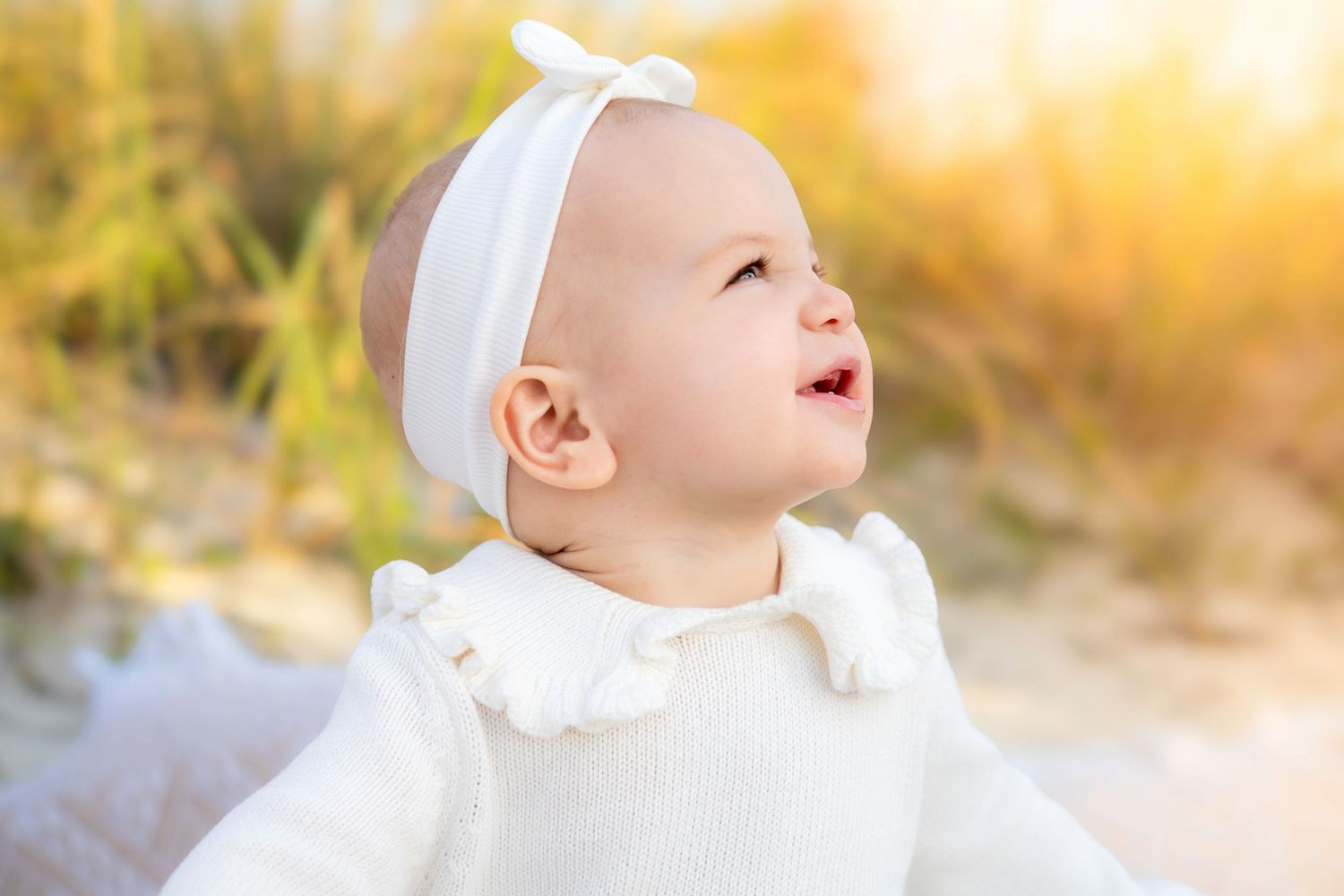 Baby in white outfit smiles while looking up during golden hour outdoor photoshoot.