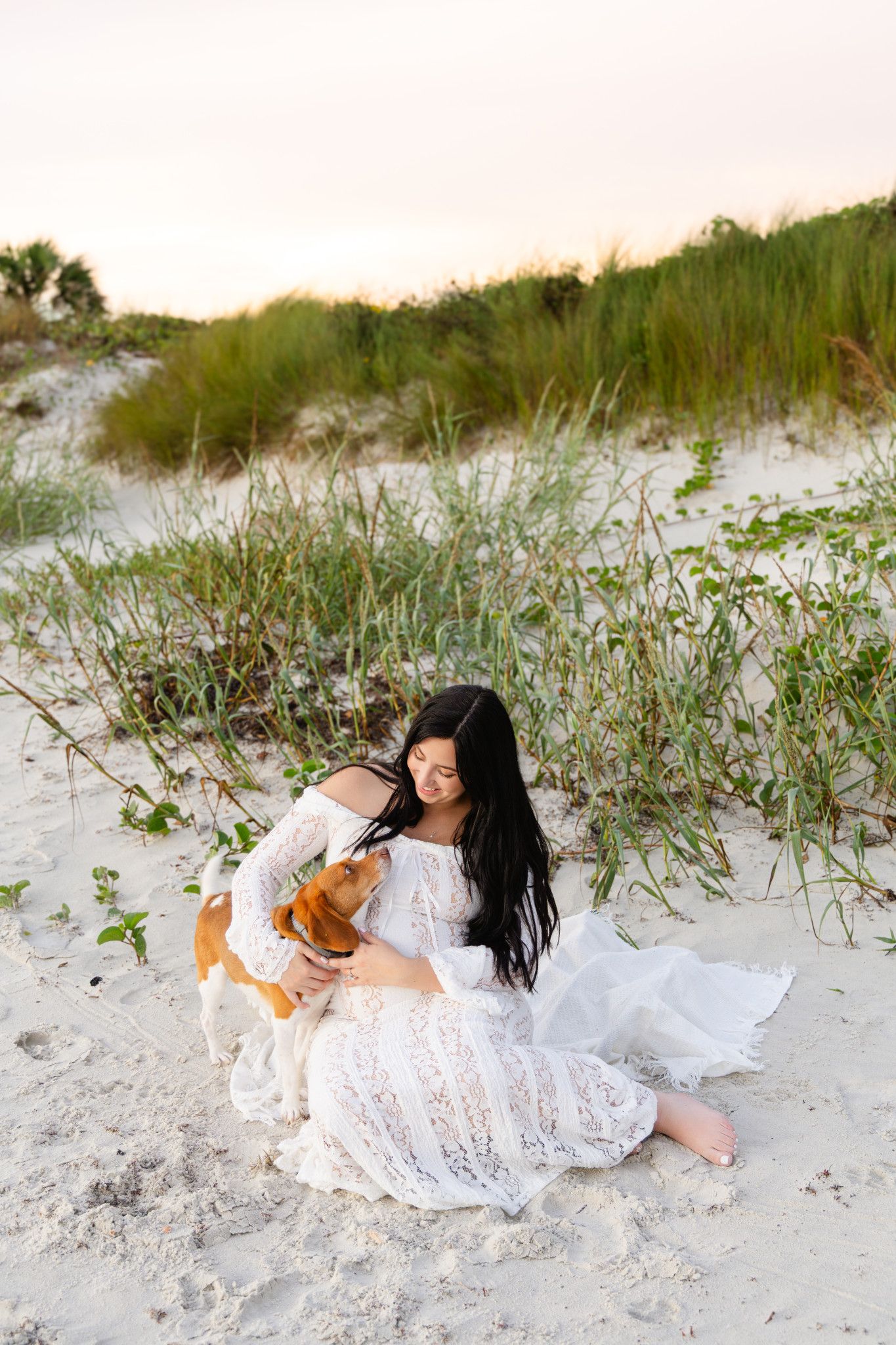 Woman in white lace dress sitting on beach sand with a Beagle dog, dunes and grass in background at sunset.