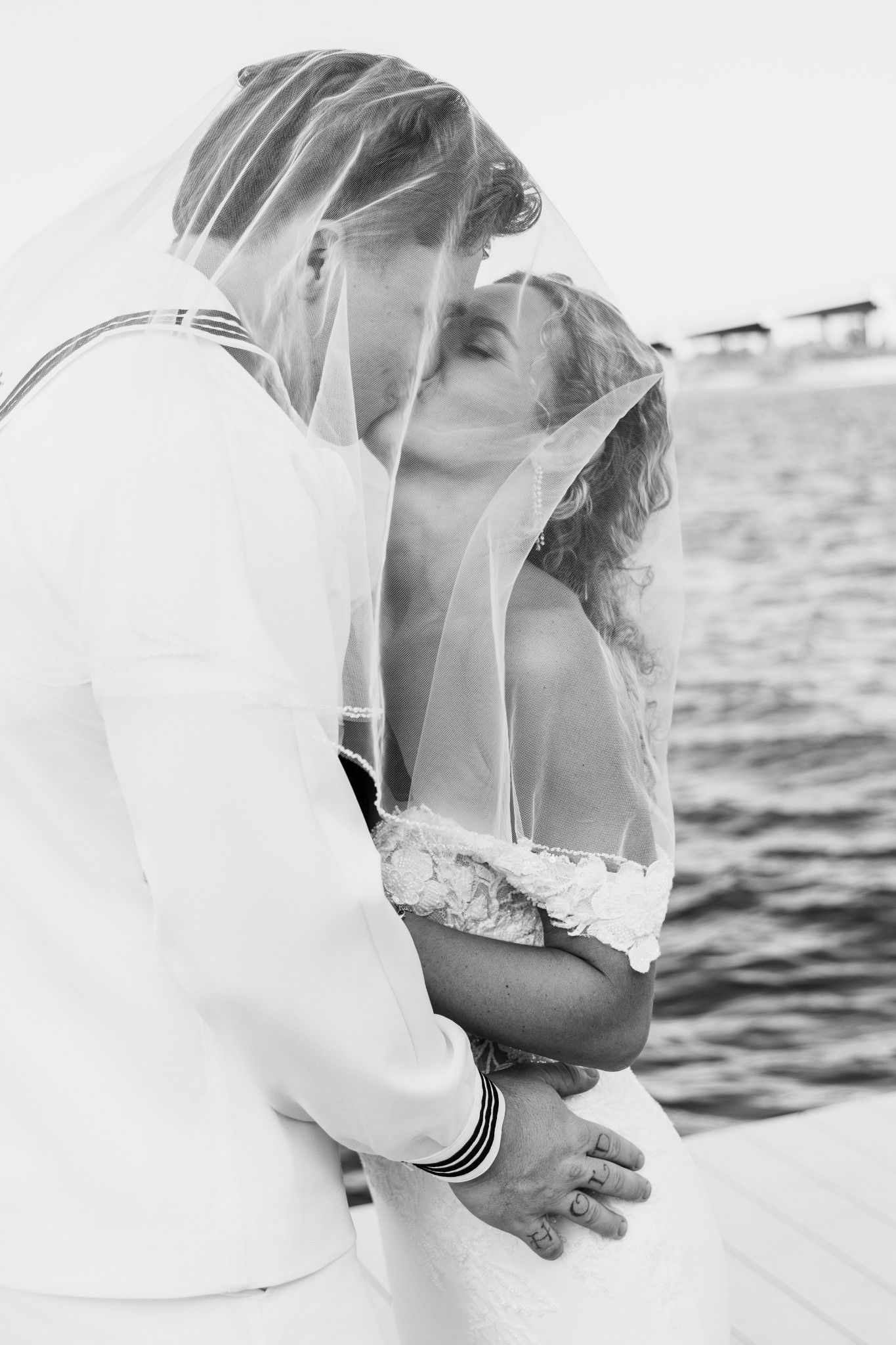 Black and white photo of a bride and groom kissing under a veil near the water on their wedding day.