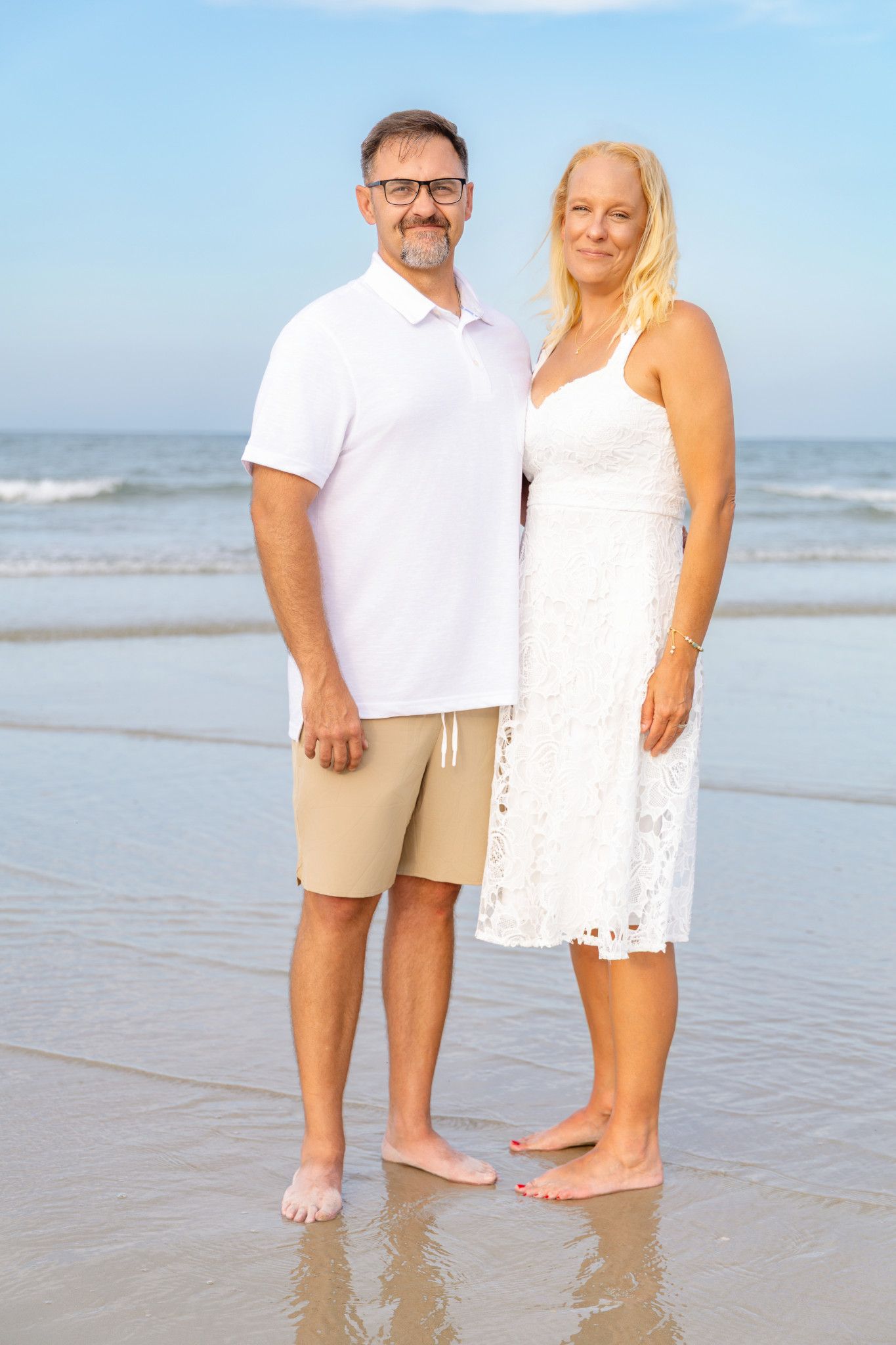 Middle-aged couple in white outfits standing barefoot on a sunny beach with ocean waves in the background.