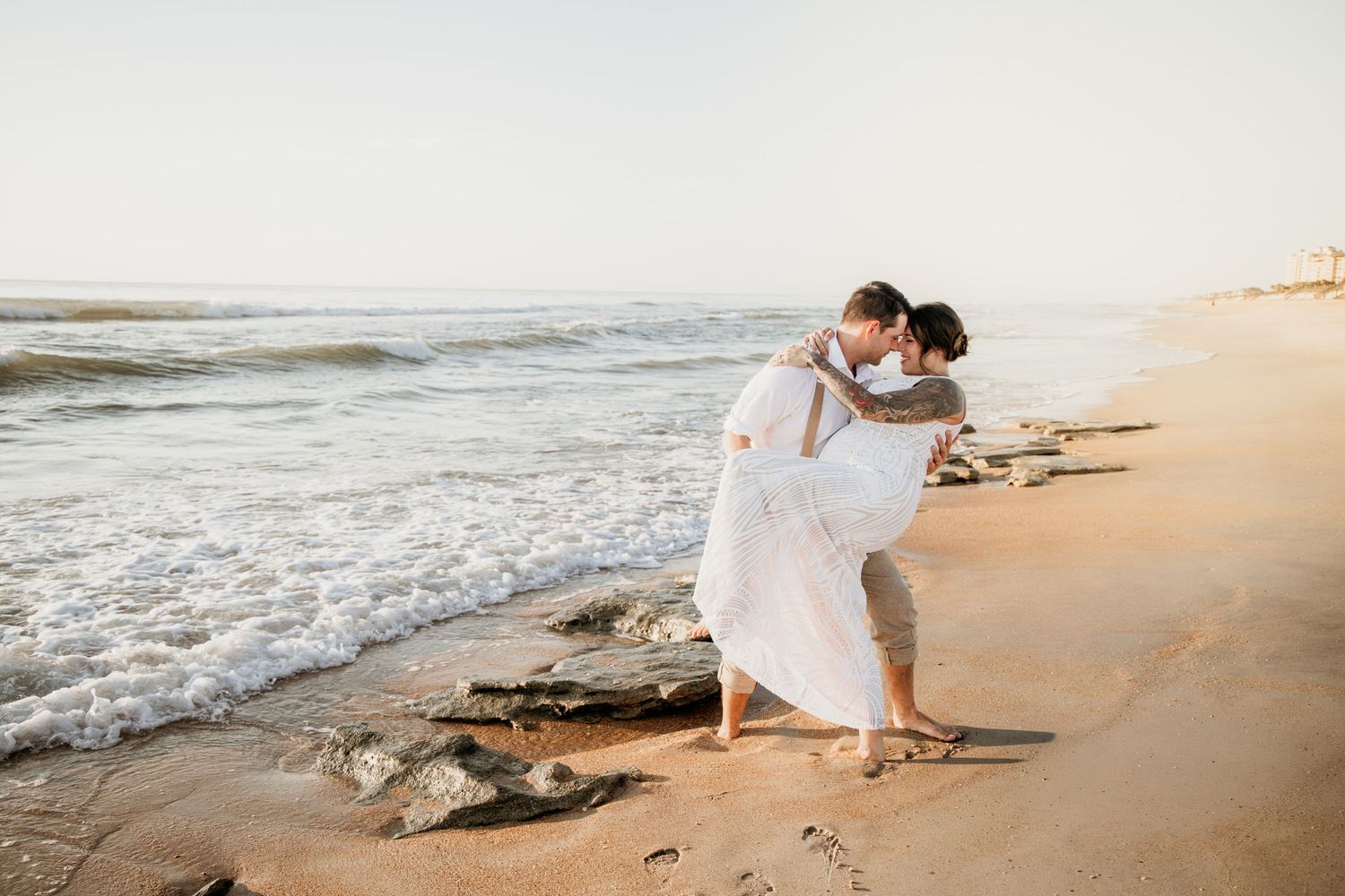 Groom lifting bride on sandy beach shoreline during romantic wedding photo session at sunset.