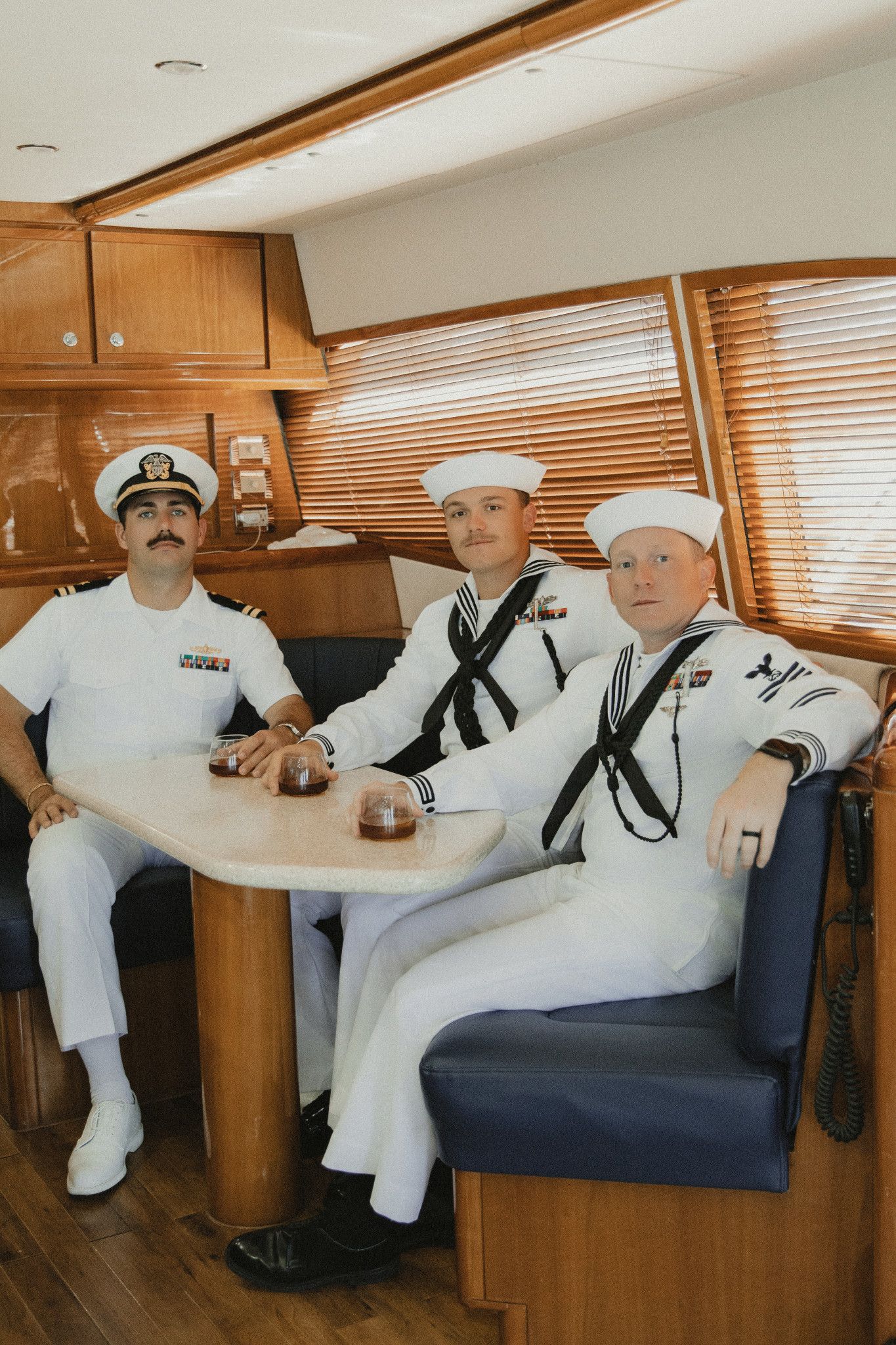 Three Navy sailors in white uniforms sit together inside a wooden boat cabin, posing for a photo at a small table.