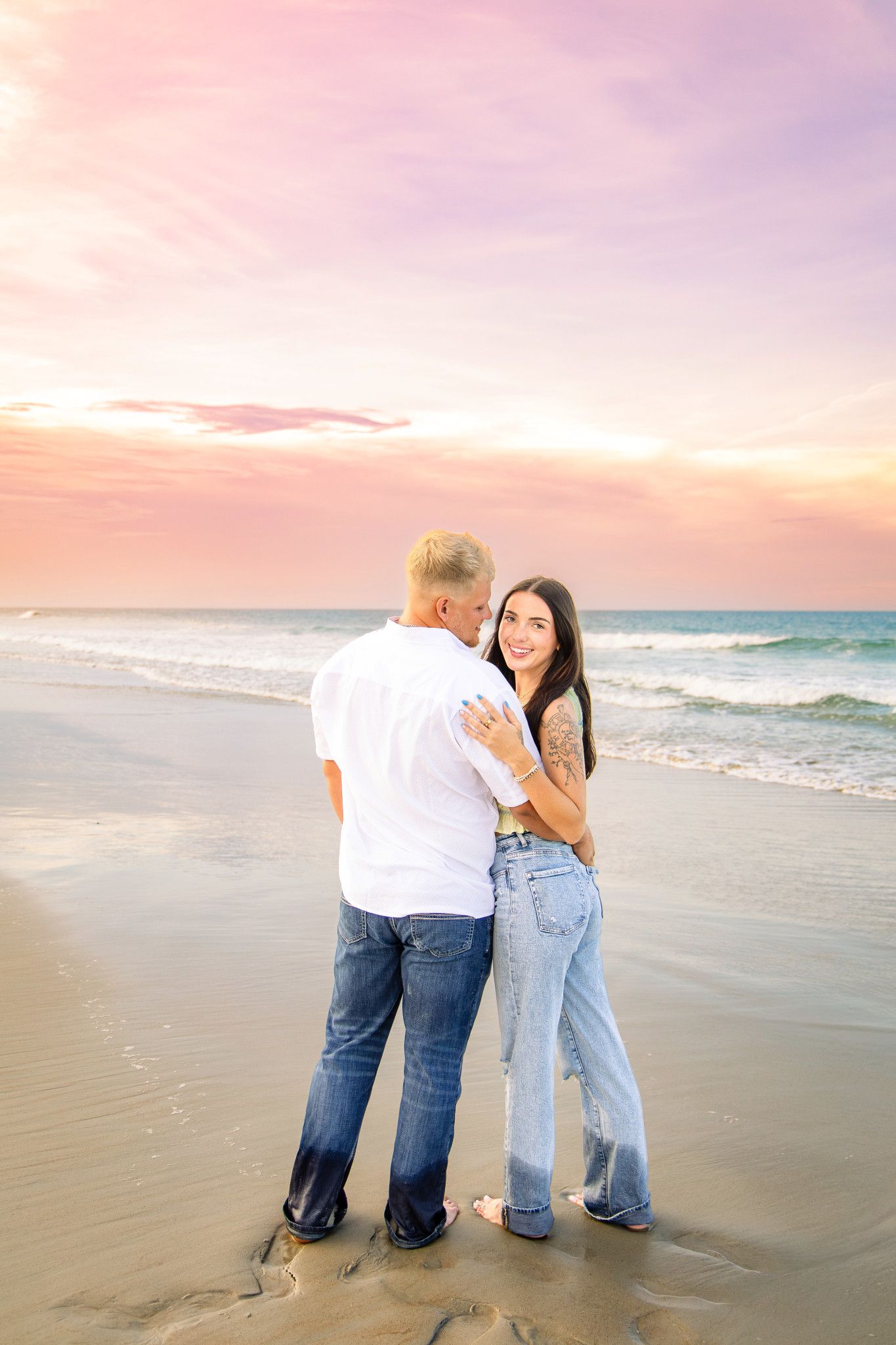 Couple embracing on a beach at sunset, woman smiling at camera, both wearing jeans, pastel pink sky backdrop.