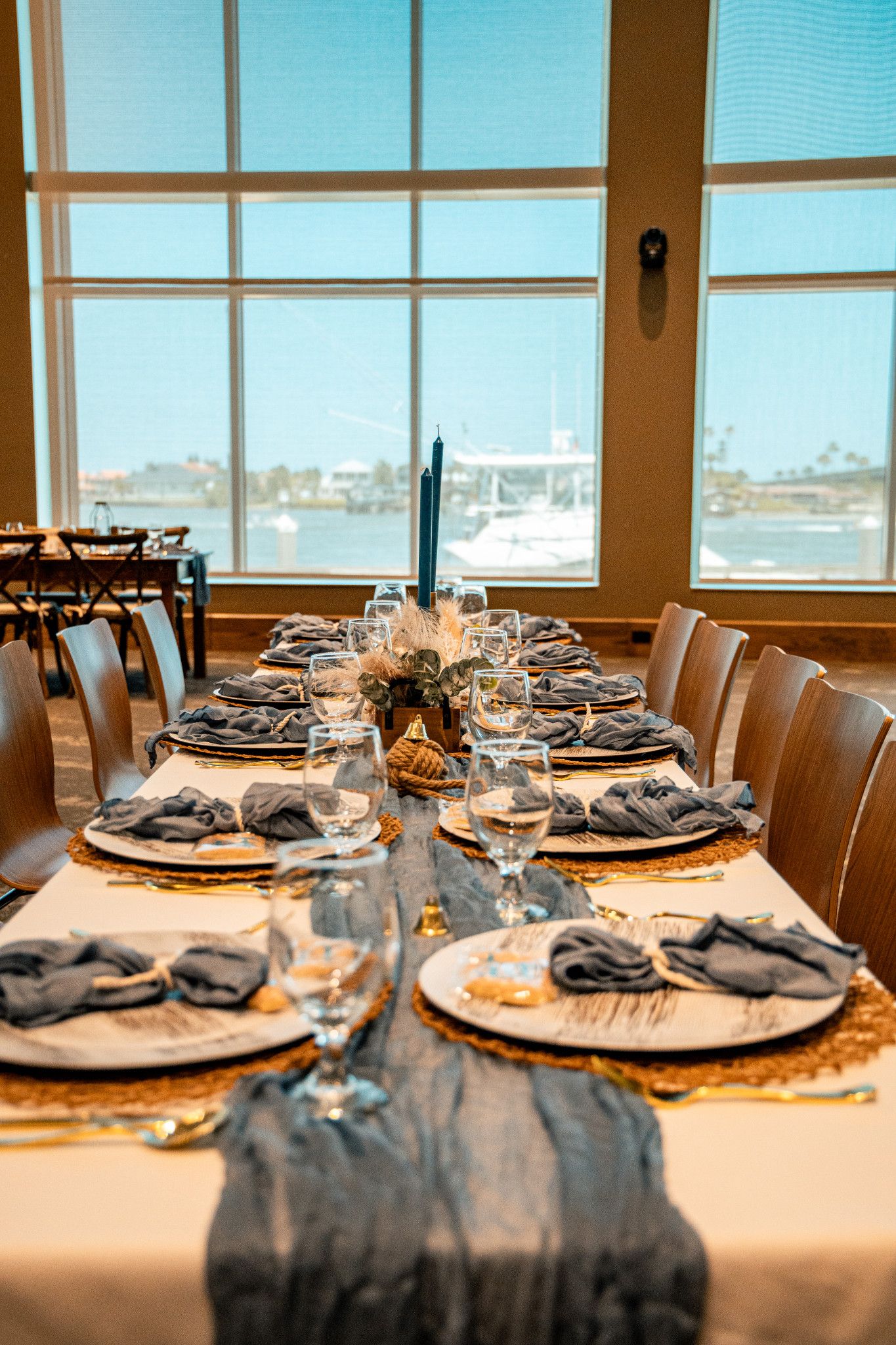 Elegant waterfront dining table set with blue napkins, gold chargers, and candles overlooking a marina.