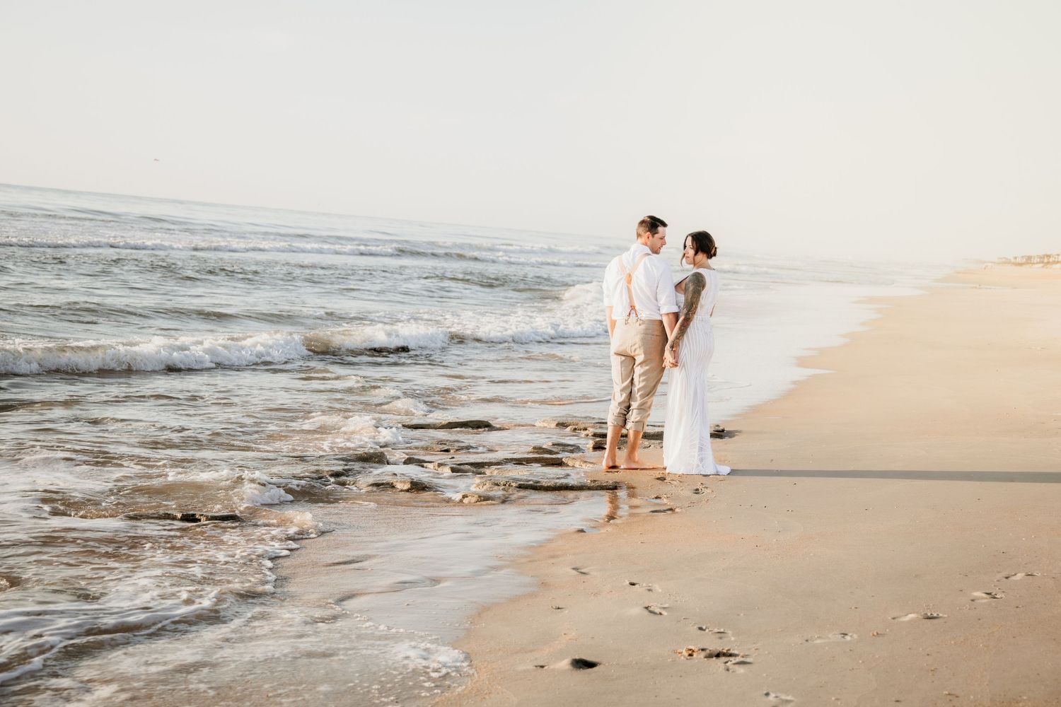 Couple in white attire standing romantically on a sandy beach shore with gentle ocean waves at golden hour.