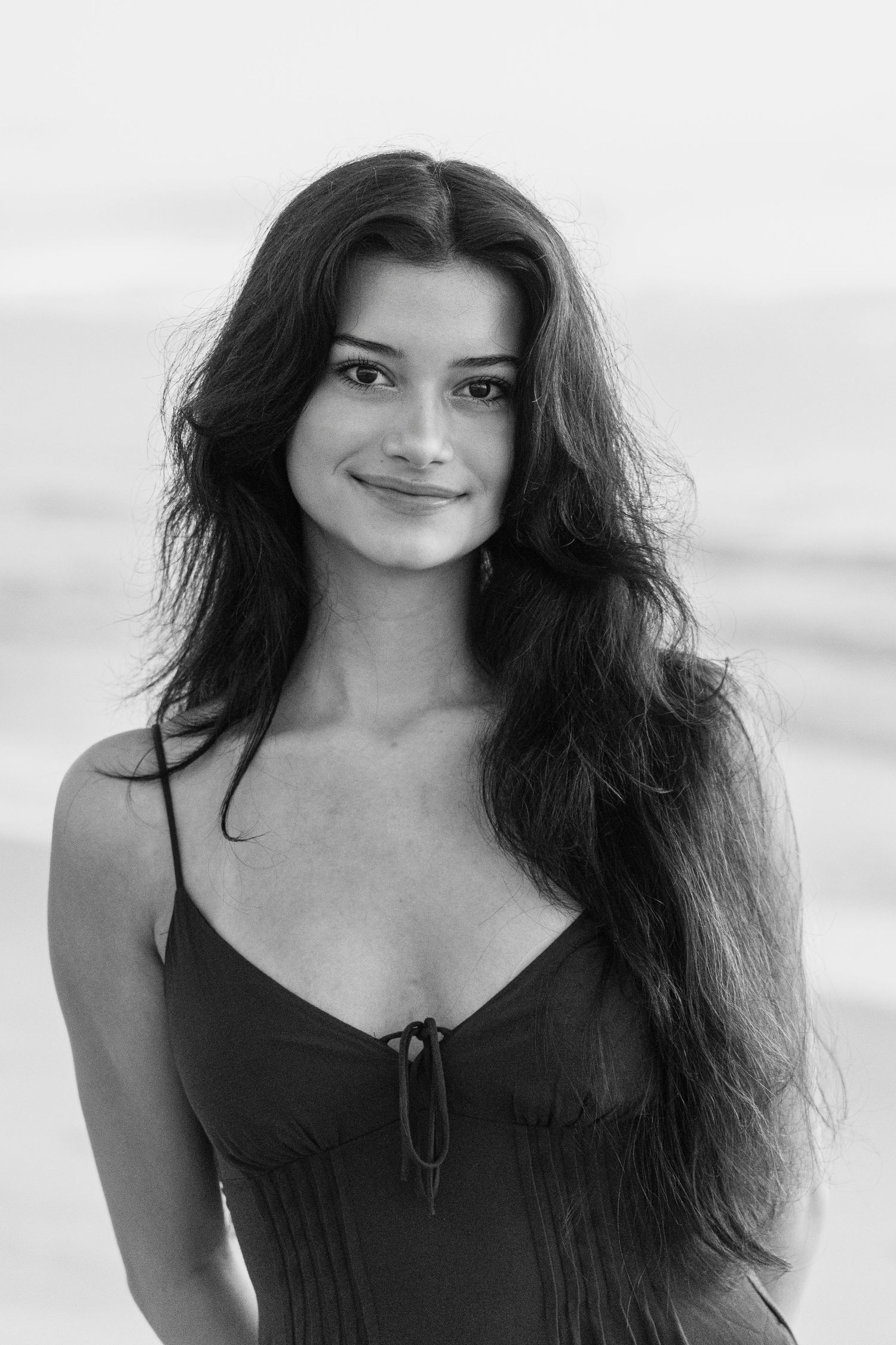 Black and white portrait of a young woman with long dark hair smiling at the beach in a spaghetti strap dress.