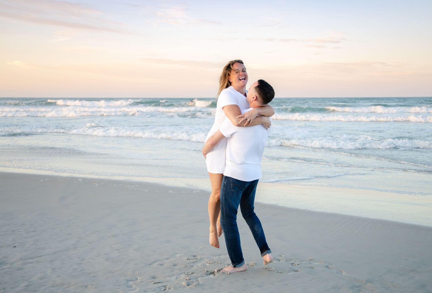 Couple embracing on a sandy beach at sunset, man lifting woman in white tops, ocean waves in background.