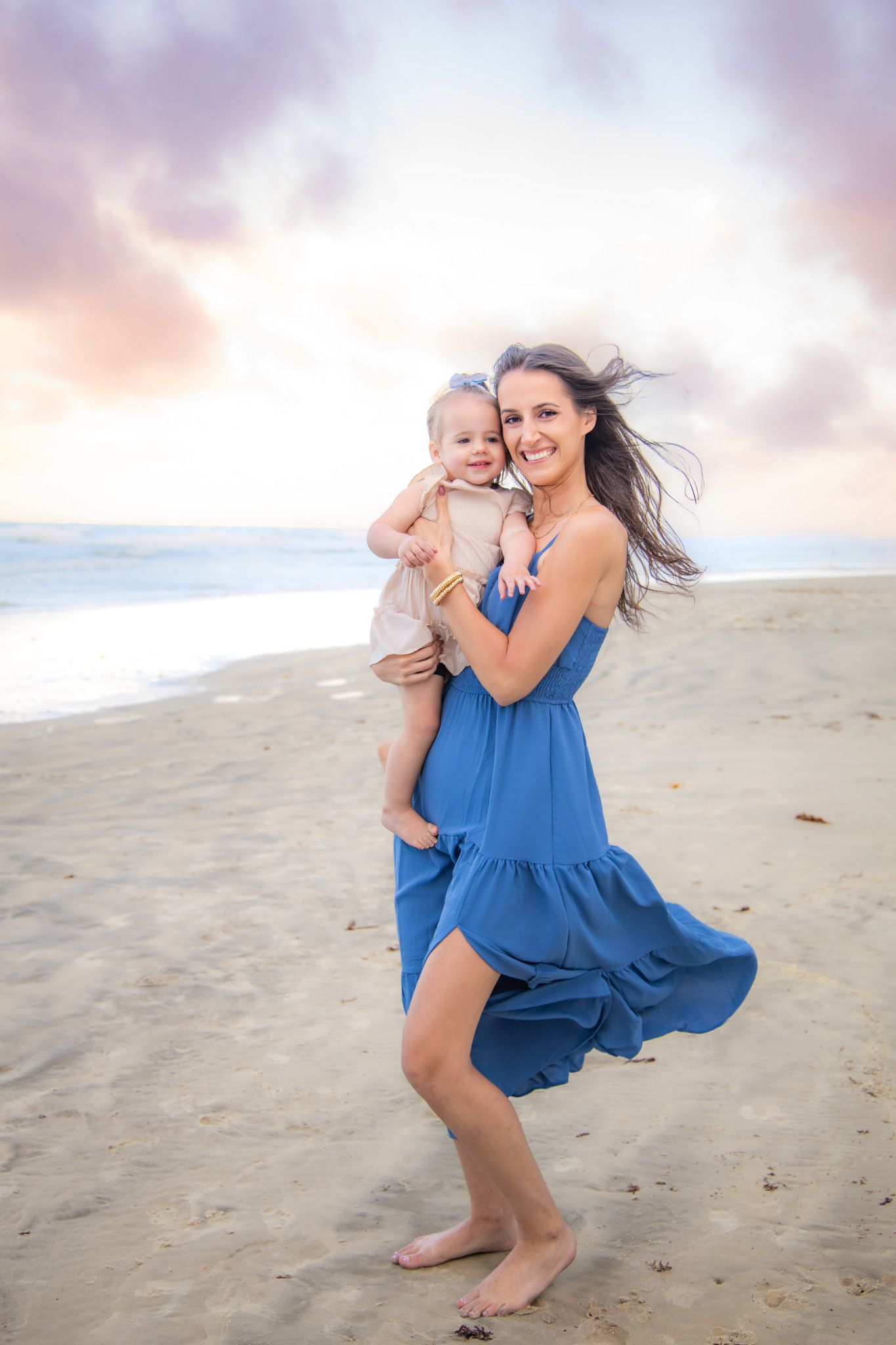 A smiling woman in a blue dress holds a baby on a sandy beach at sunset with colorful clouds in the background.