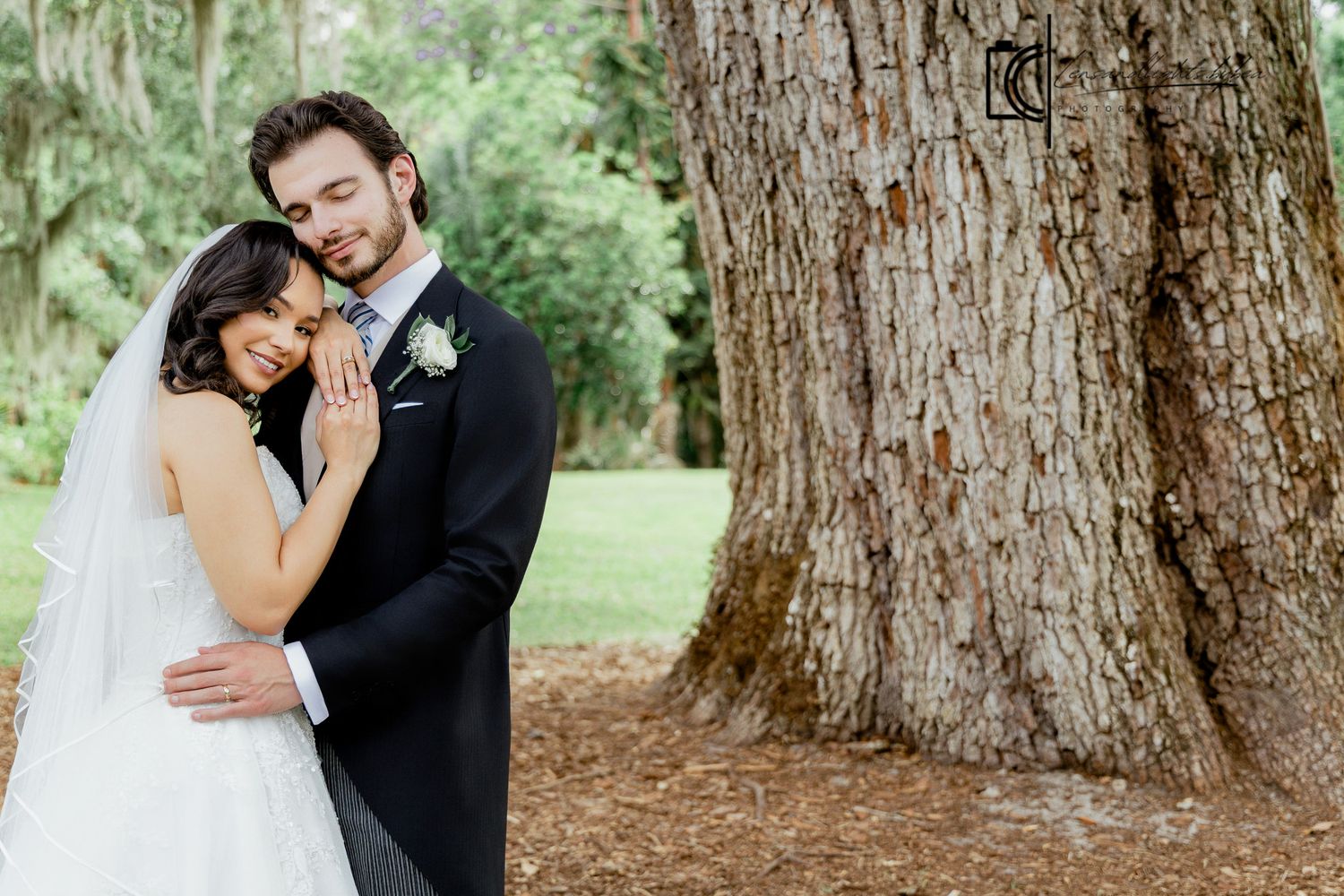Bride and groom embrace romantically beside a large tree trunk outdoors, bride in white gown with veil, groom in navy suit.