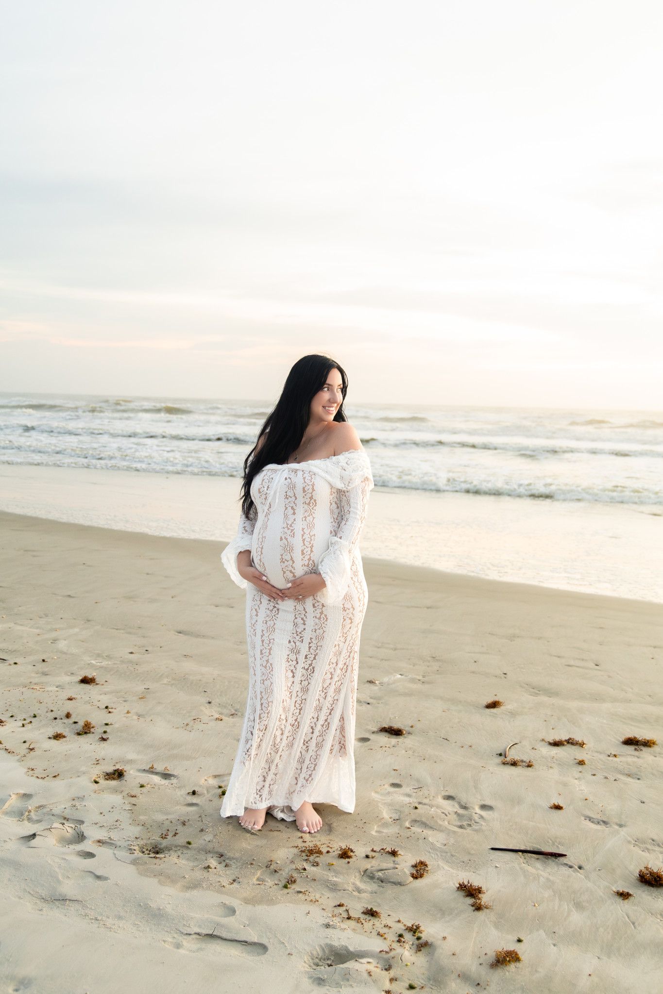 Pregnant woman in white lace off-shoulder gown standing on sandy beach at sunset, cradling baby bump.