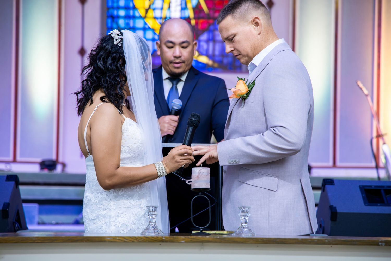 Groom placing wedding ring on bride's finger during church ceremony, officiant standing behind them.