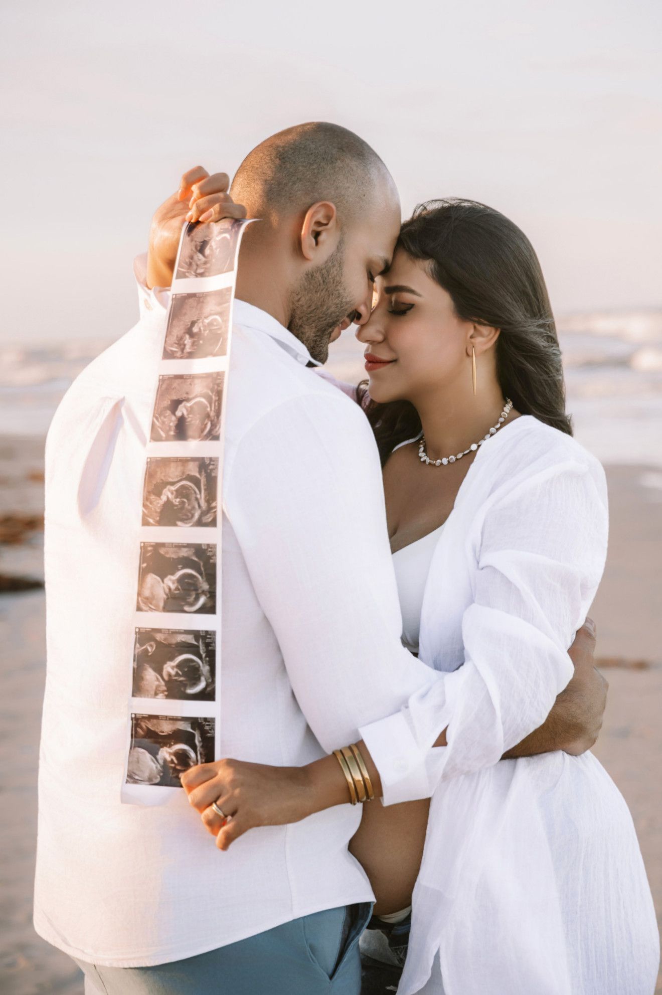 Couple in white outfits sharing a tender moment on beach, holding ultrasound photos, announcing pregnancy.