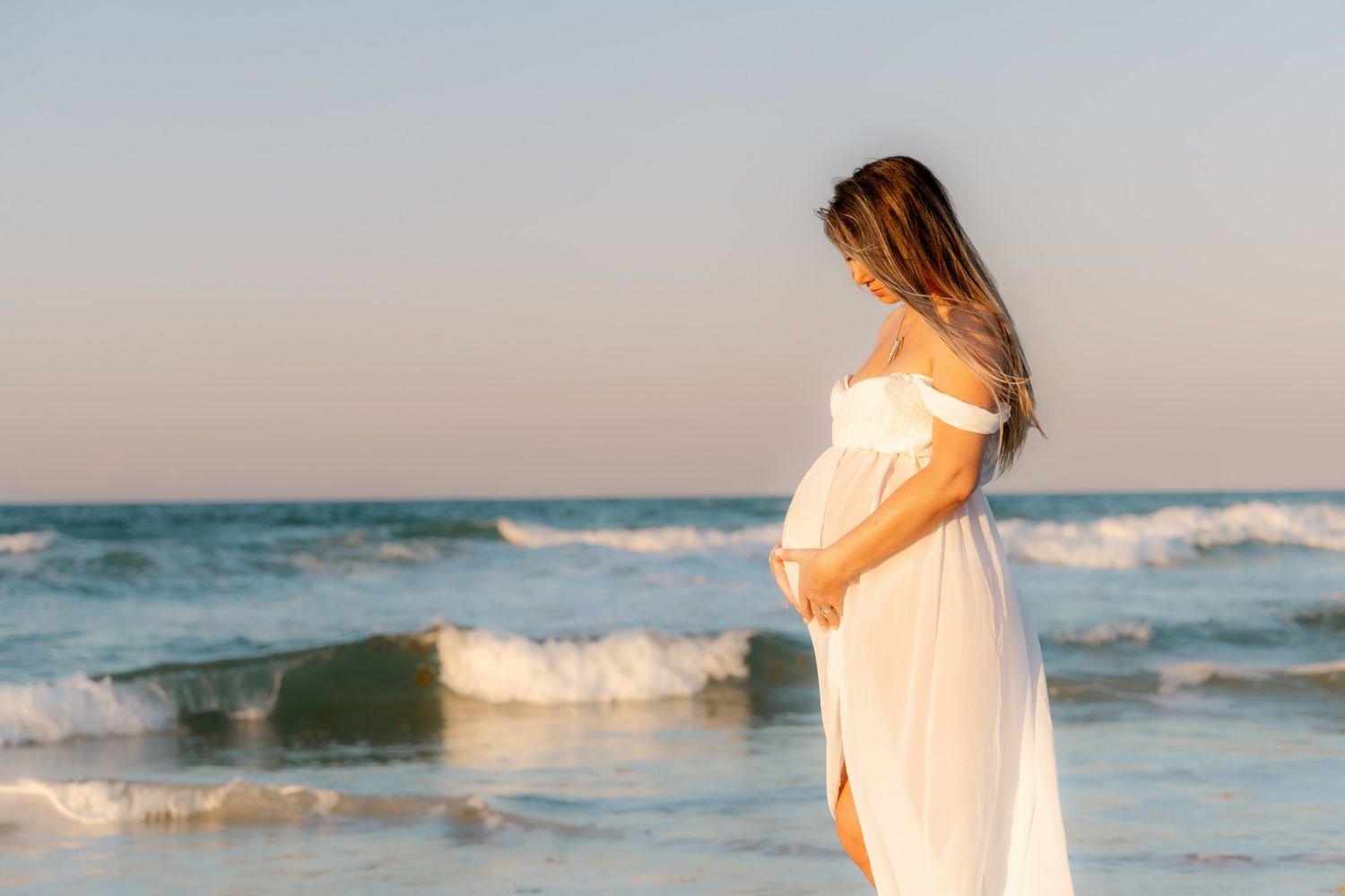 Pregnant woman in white dress standing on beach at sunset, cradling her baby bump near ocean waves.