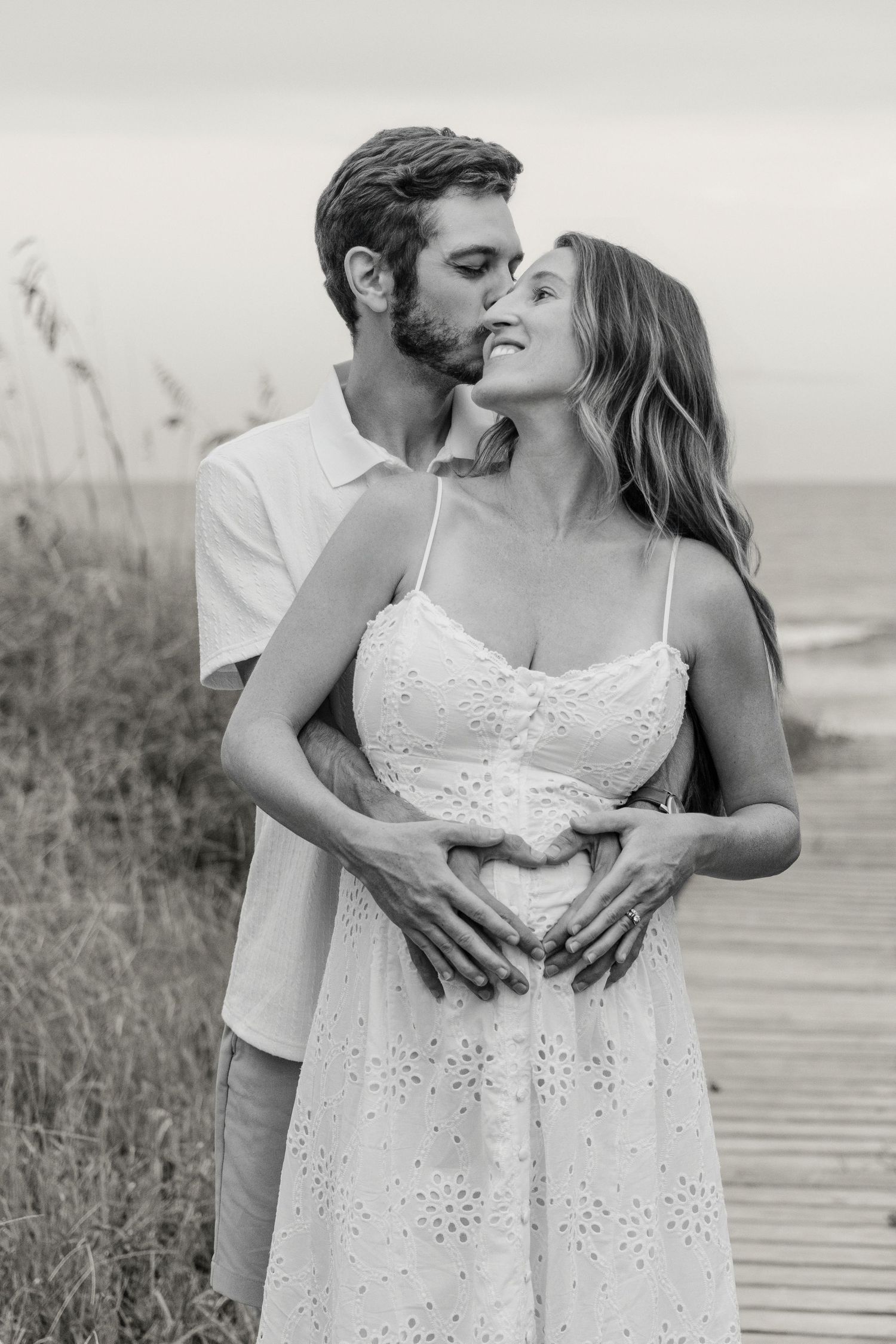 Black and white photo of a couple on a beach boardwalk, man kissing woman's cheek as they form a heart on her pregnant belly.