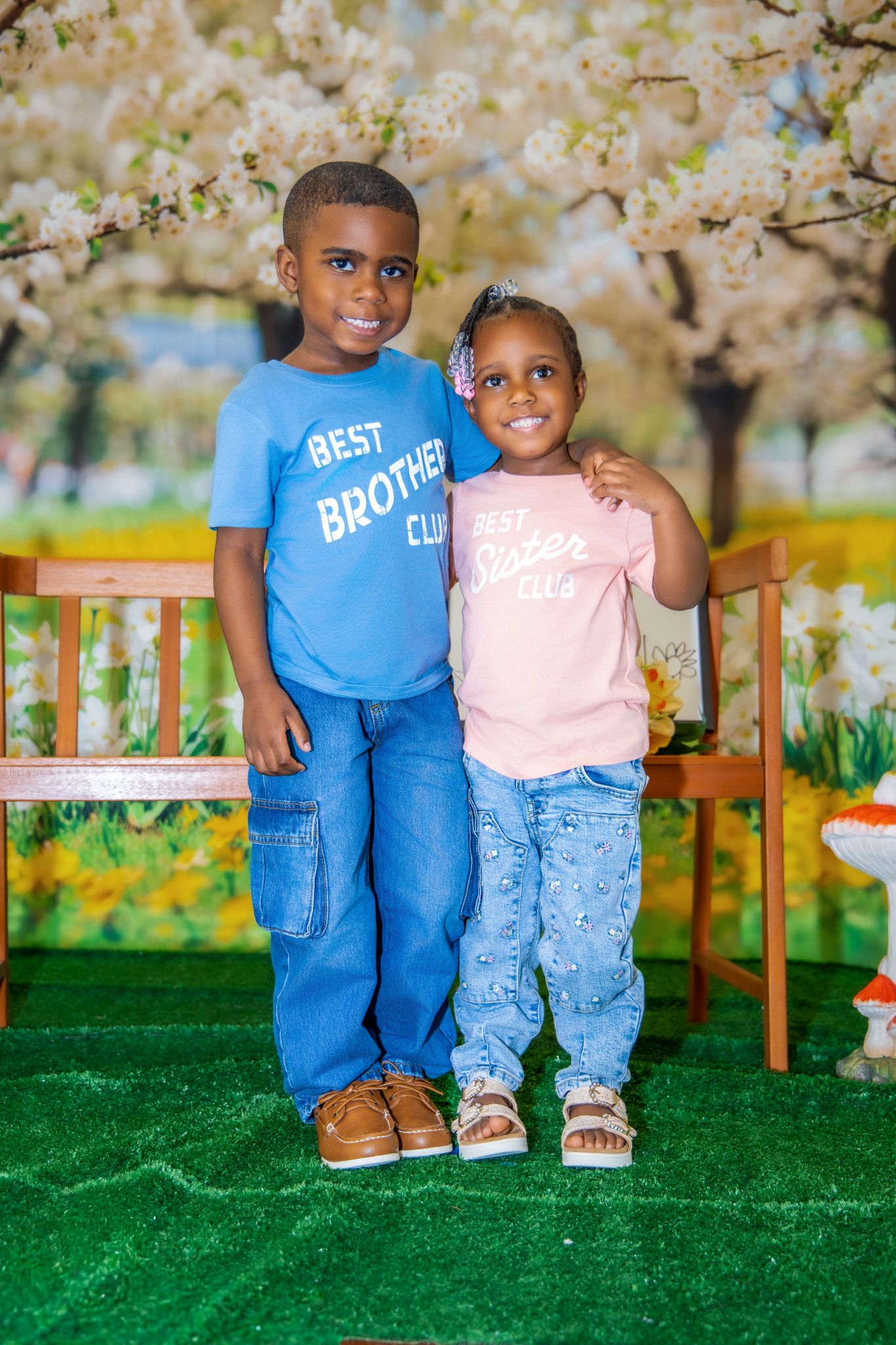 Two young children in blue and pink outfits pose together against a spring cherry blossom backdrop on green grass.