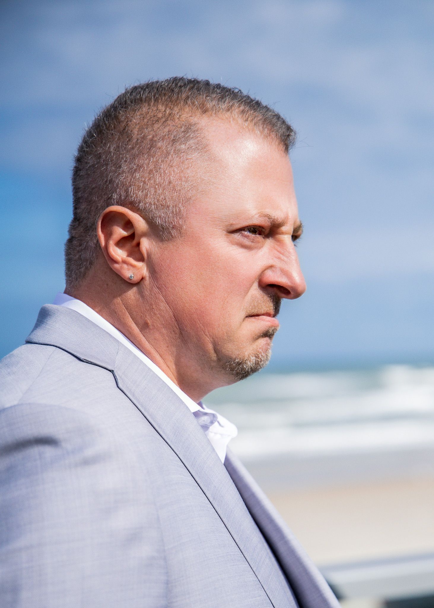 A person in a light gray suit looks to the side while standing on a sunny beach with ocean waves in the background.