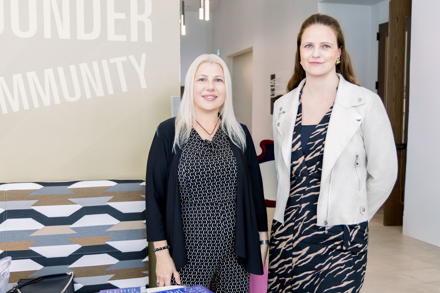 Two professional women smiling and standing together near a community center sign at a business event.