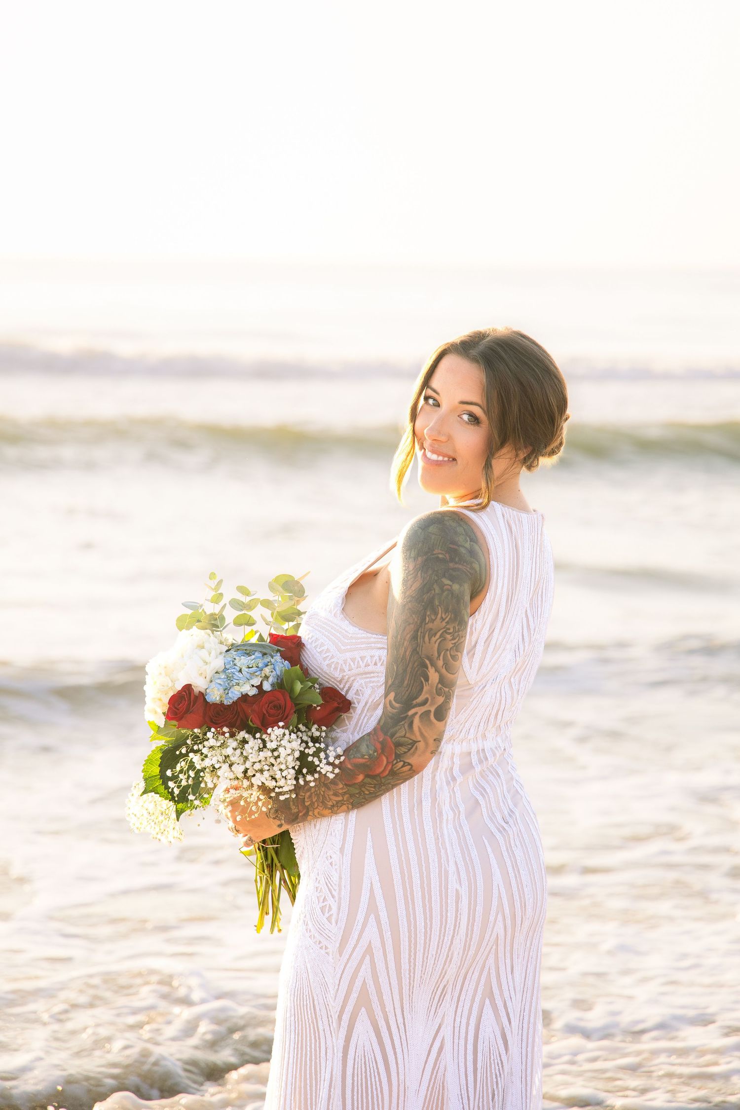 Tattooed bride in white dress holding colorful bouquet on a sunlit beach, smiling over shoulder.