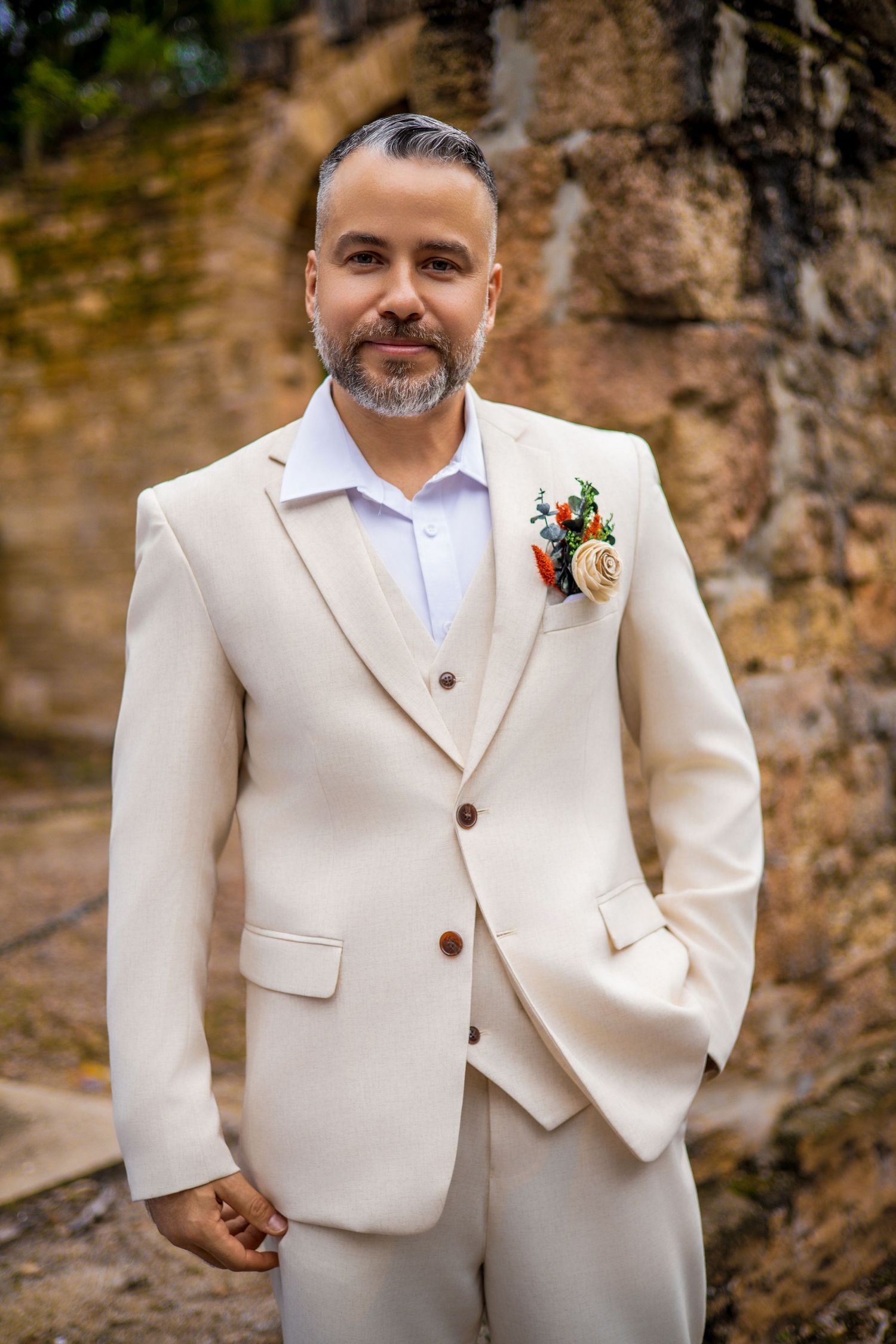 A person wearing a cream colored suit stands confidently against a stone wall.