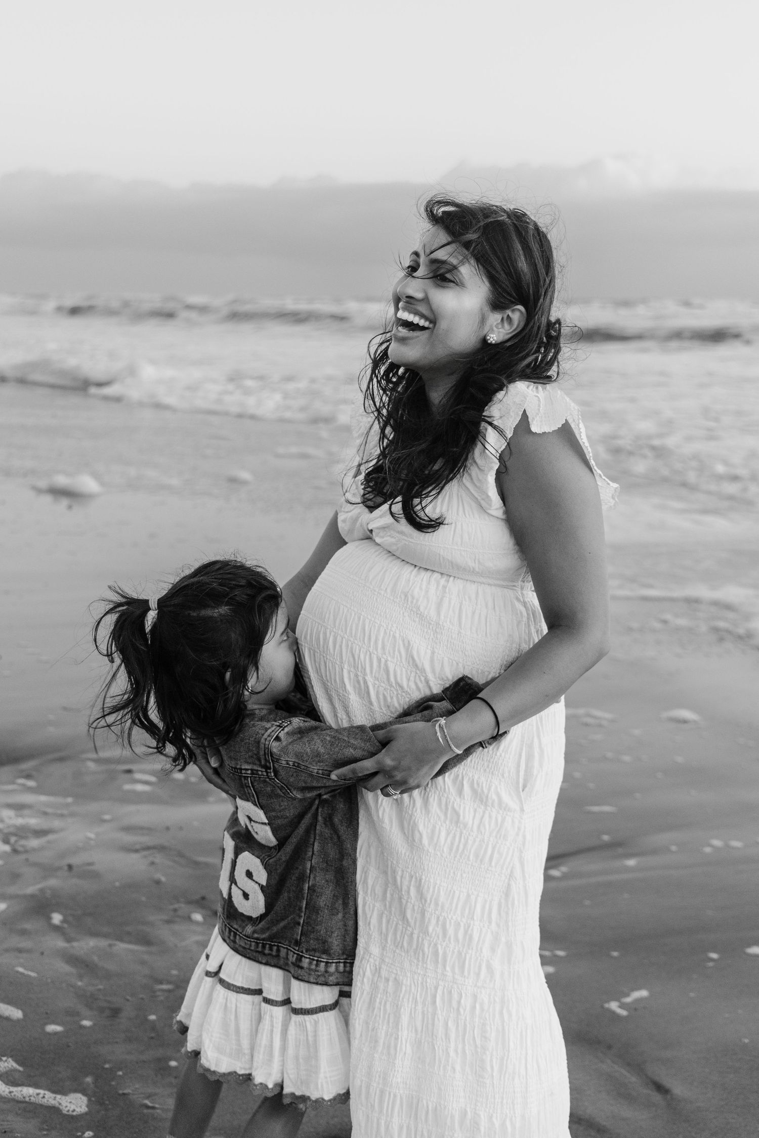 Black and white photo of a smiling pregnant woman in a white dress embraced by her young daughter on the beach.