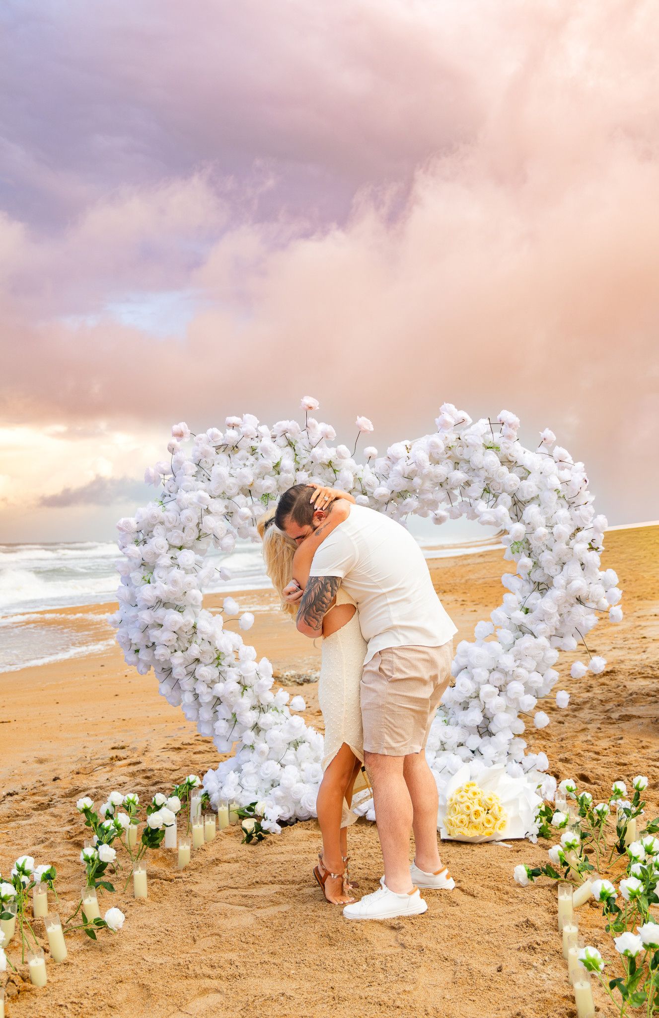 Couple embracing on a sandy beach inside a heart-shaped white flower arch with candles and rose petals at a colorful pink sunset.