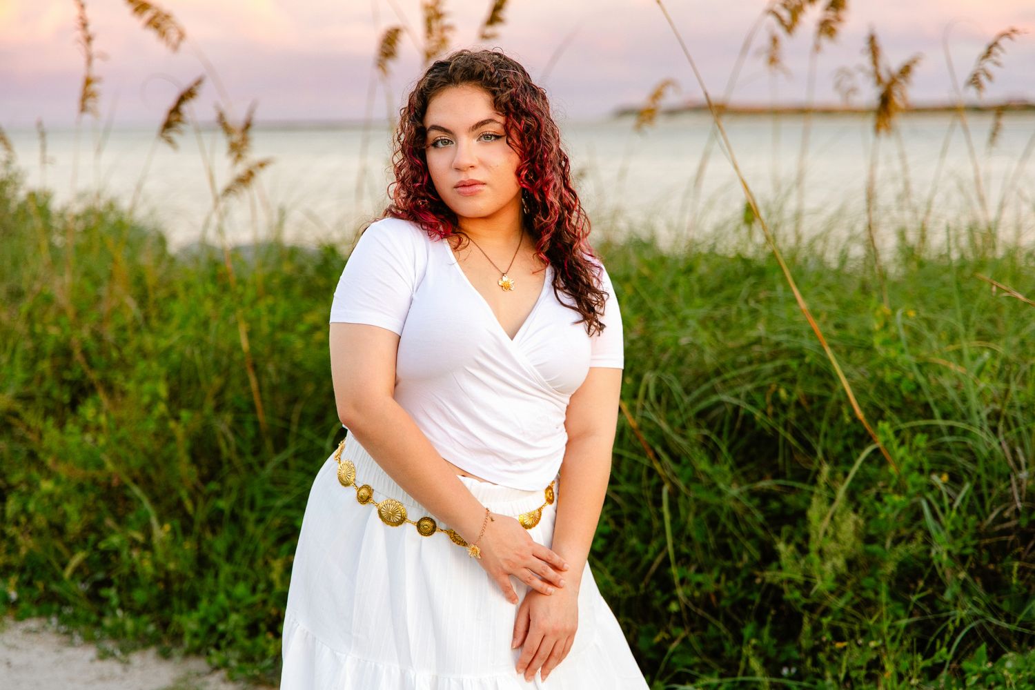 Young woman with long curly hair in a white dress with a gold belt standing by tall beach grass at sunset near the water.