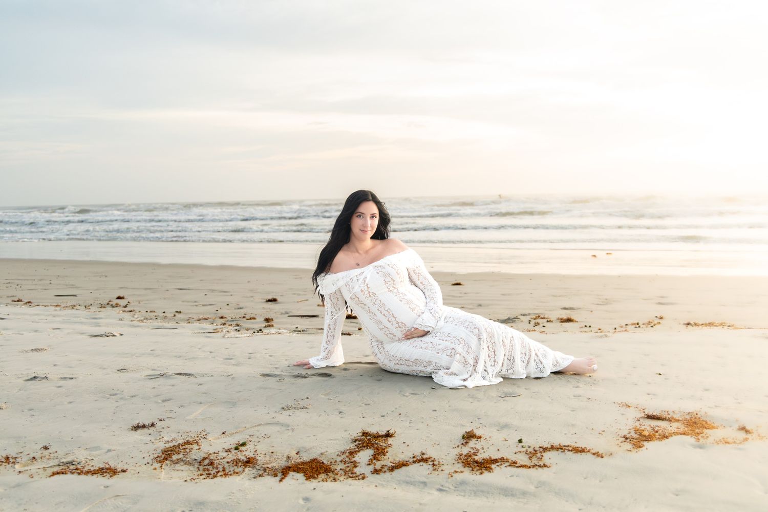 Pregnant woman in white off-shoulder gown sitting on sandy beach at sunset with ocean waves behind her.