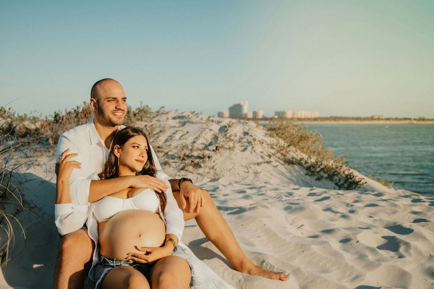 Pregnant couple sitting on sandy beach, man embracing woman with baby bump, ocean and city skyline in background.