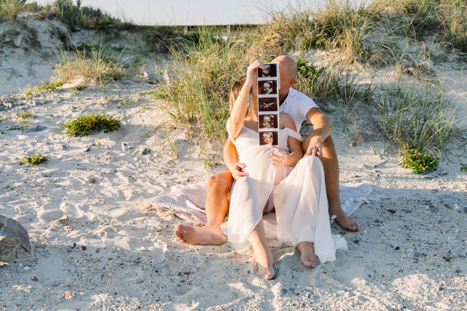Couple sitting on beach sand holding ultrasound photos in front of their faces during maternity shoot.