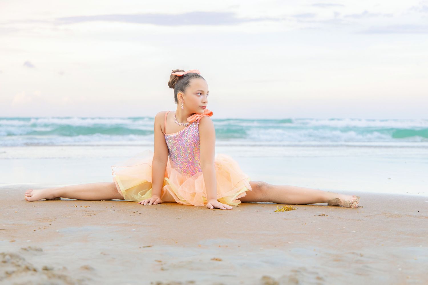 Young ballerina in pink tutu performs splits on sandy beach with ocean waves in background.