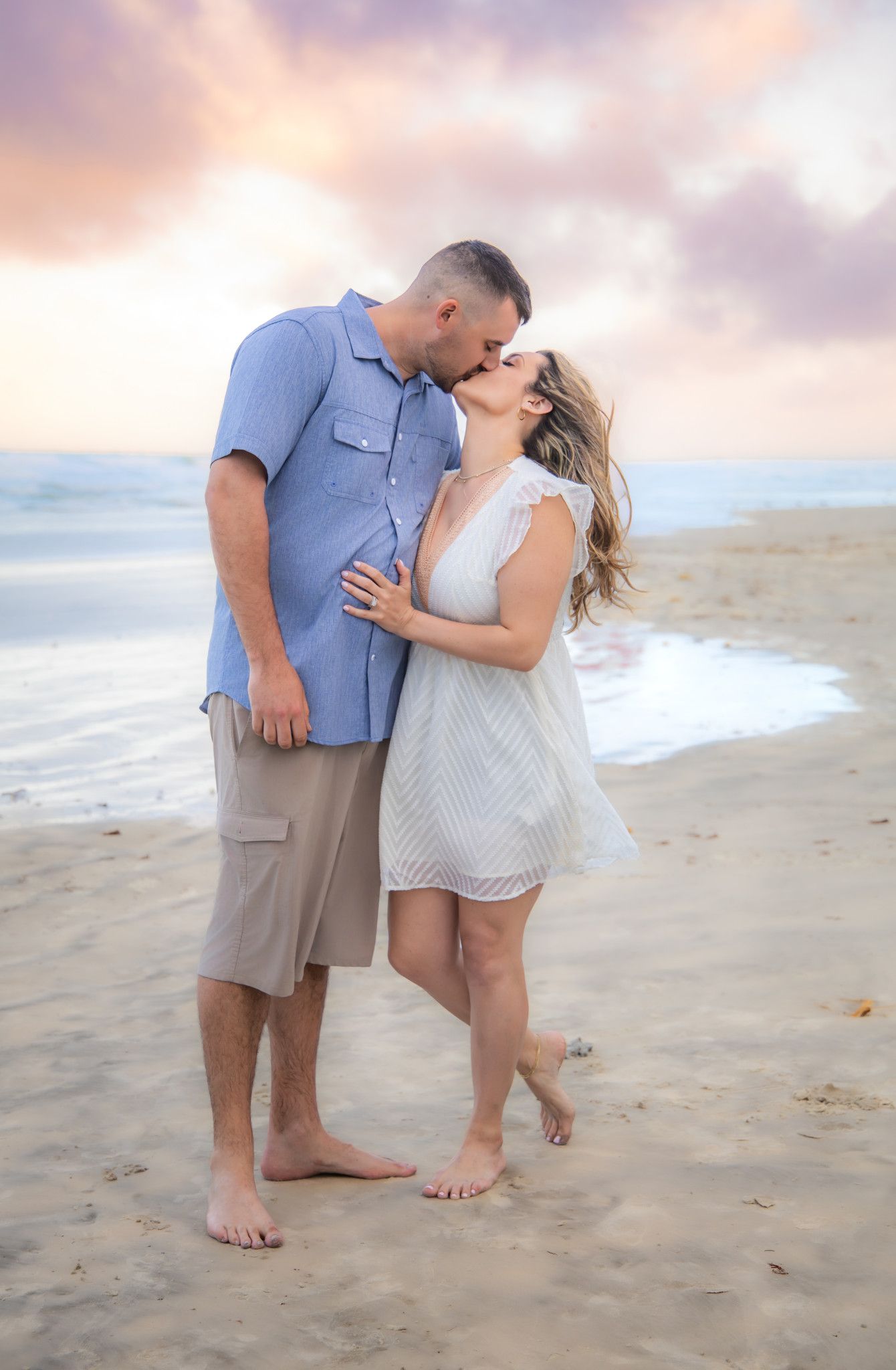 Couple sharing a romantic kiss on a sandy beach at sunset, man in blue shirt and woman in white dress.