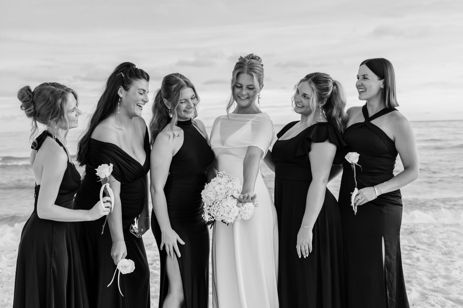 Bride in white gown with bridesmaids in black dresses laughing together on a beach in black and white photo.
