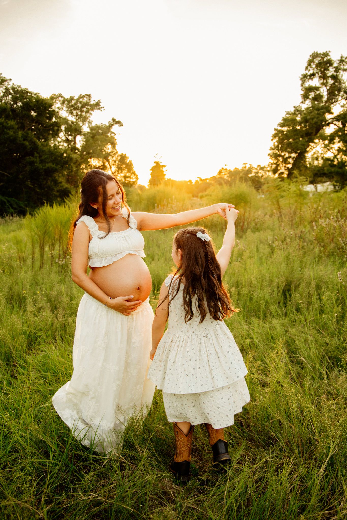 Pregnant mom in white dress twirling young daughter in cowboy boots at golden sunset.