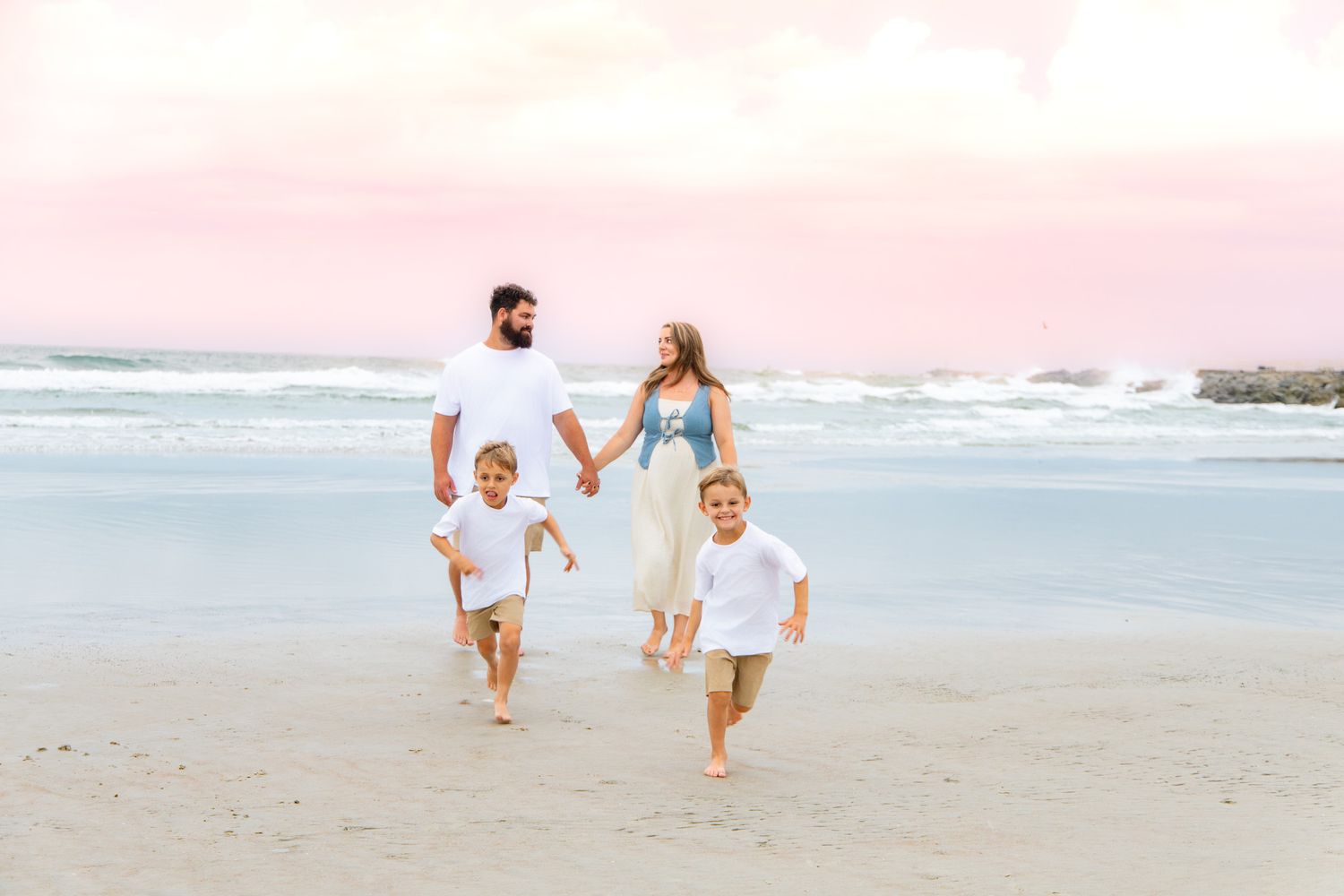 Family of four walking hand-in-hand on a beach at sunset with pink sky and ocean waves.