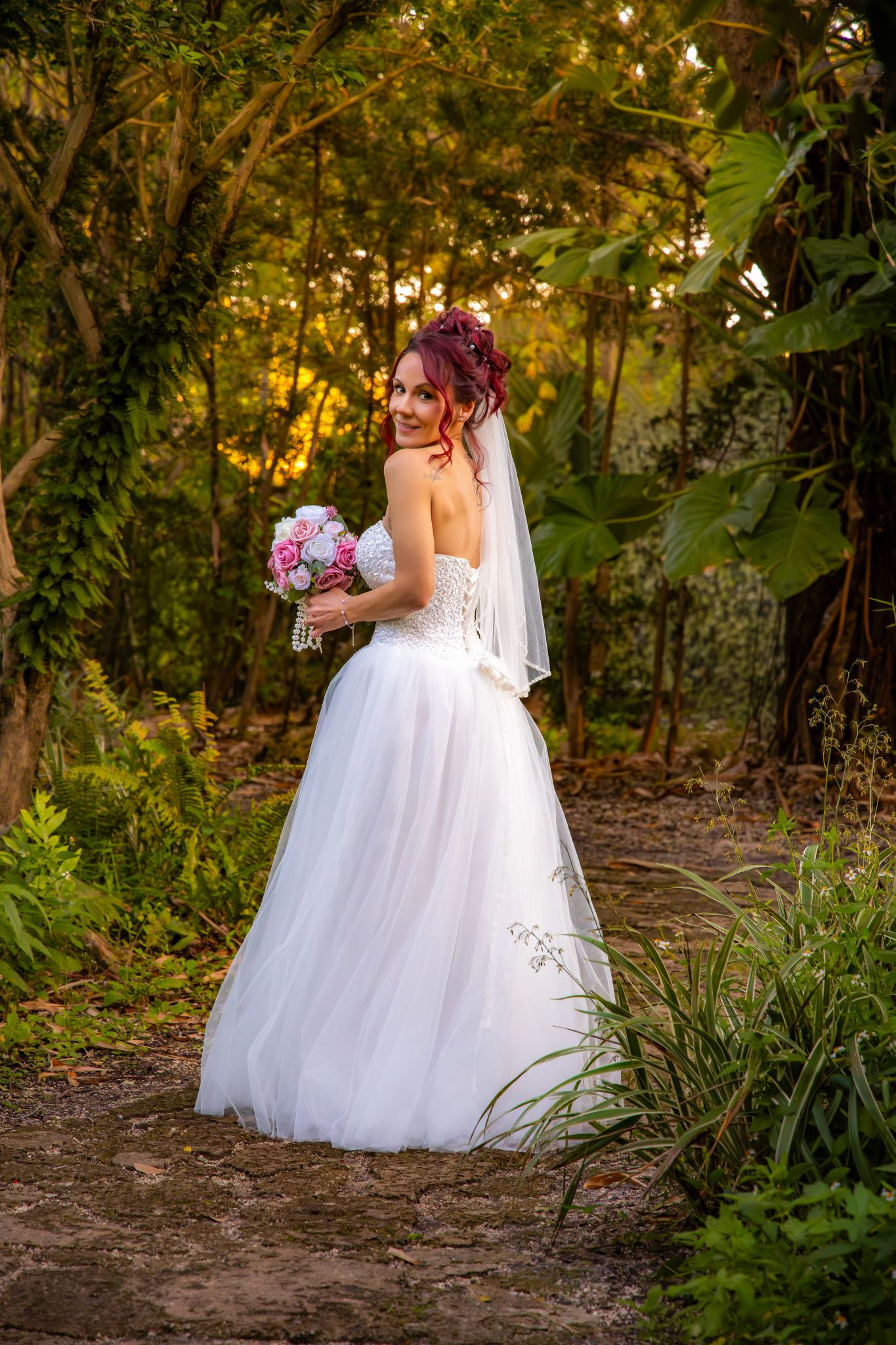 A bride in a flowing white ballgown poses in a sunlit wooded area during golden hour.