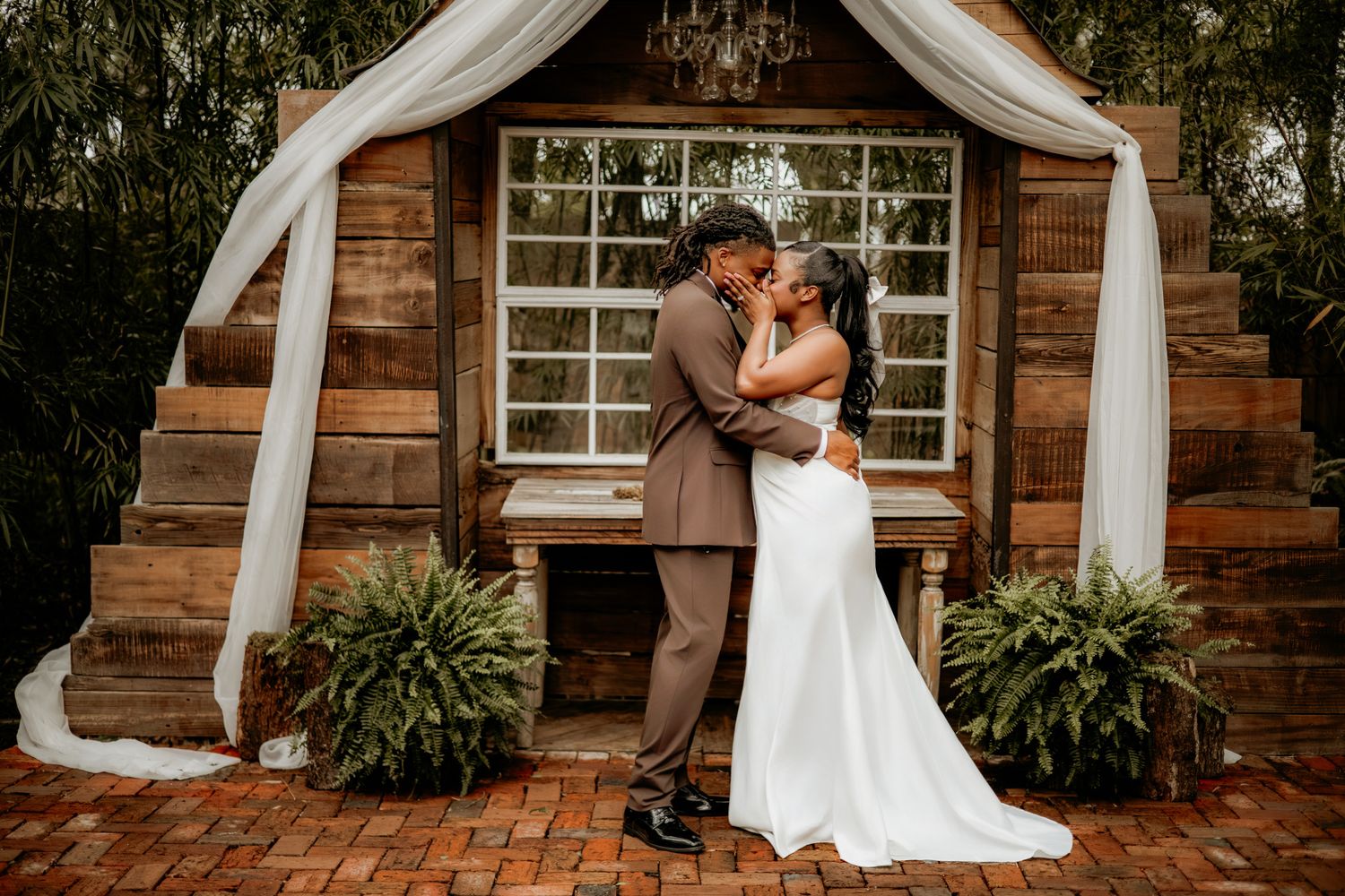 A romantic wedding couple embraces in front of a rustic wooden cabin with white drapes and greenery.