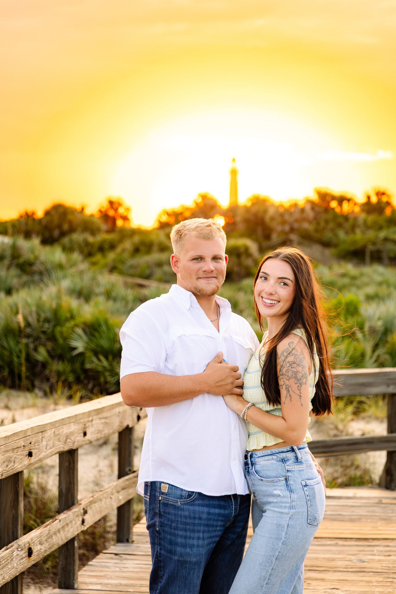 Couple embracing on a wooden boardwalk at golden sunset with a lighthouse and palm trees in the background.