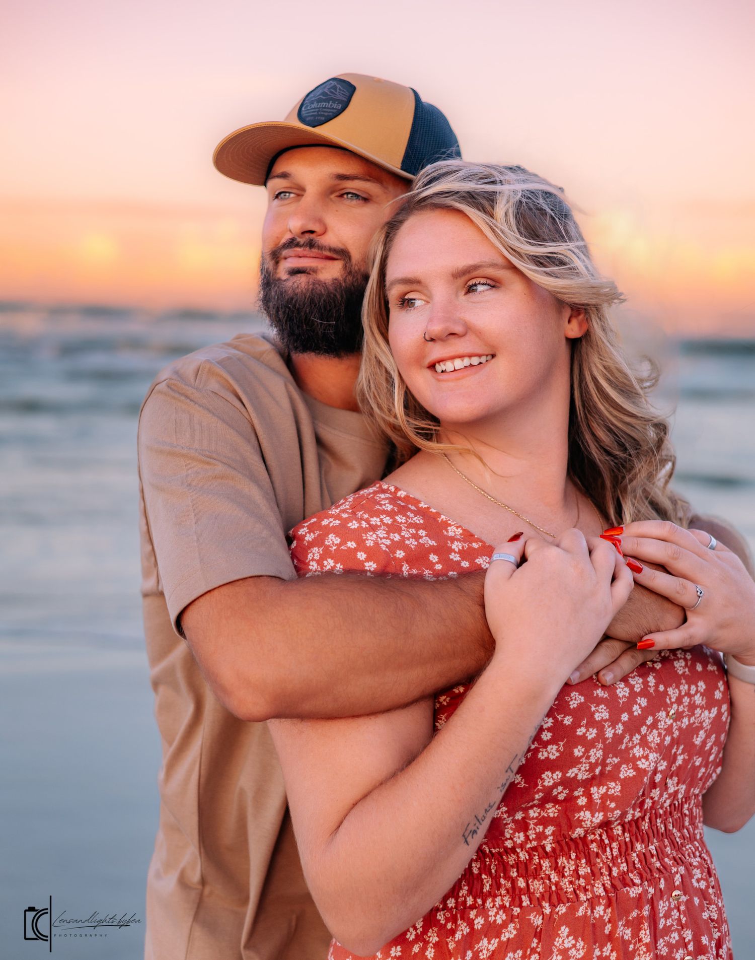 A couple embraces on the beach during sunset while wearing casual attire and a sun hat.