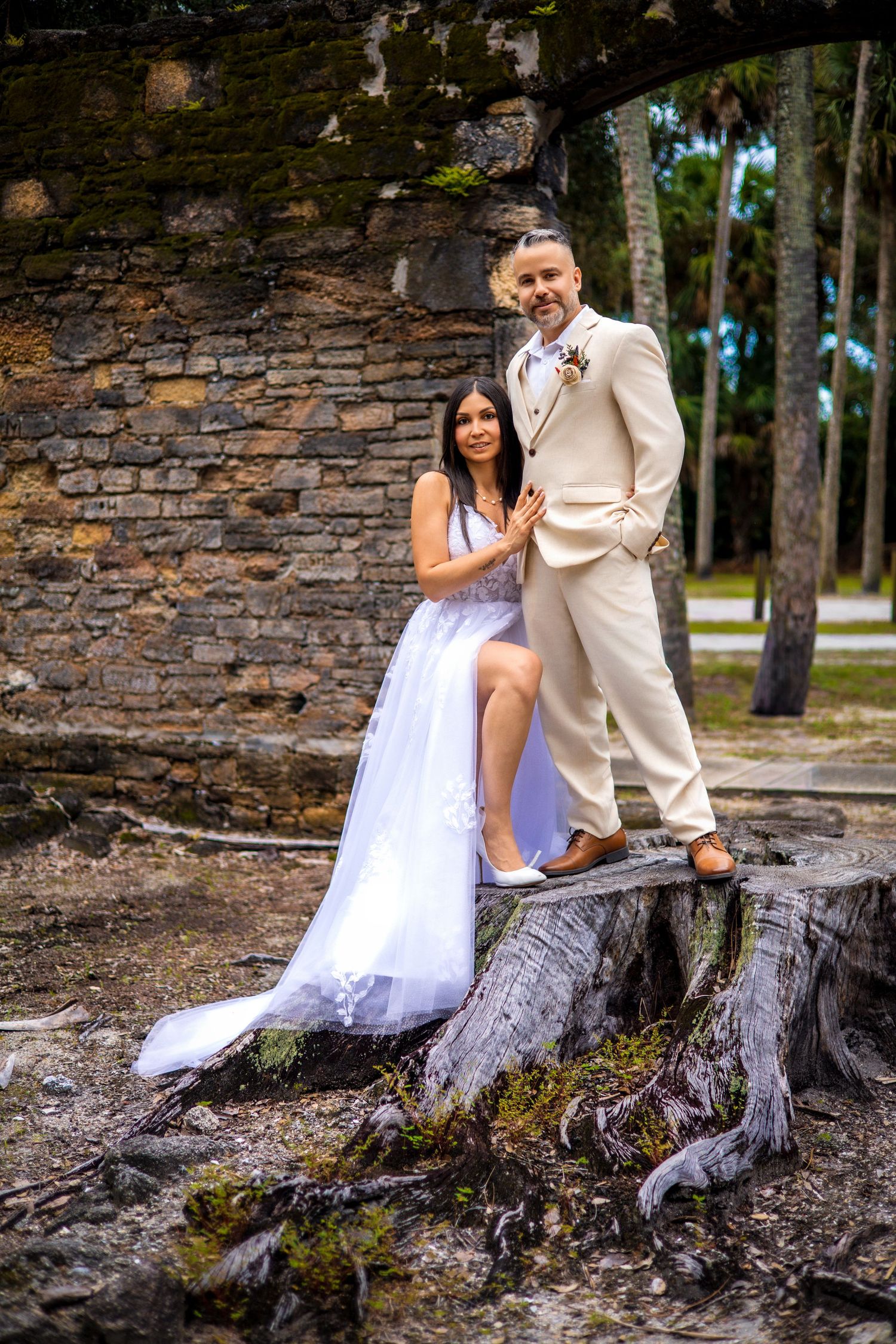 A couple in wedding attire poses together on rustic tree roots by a stone wall.