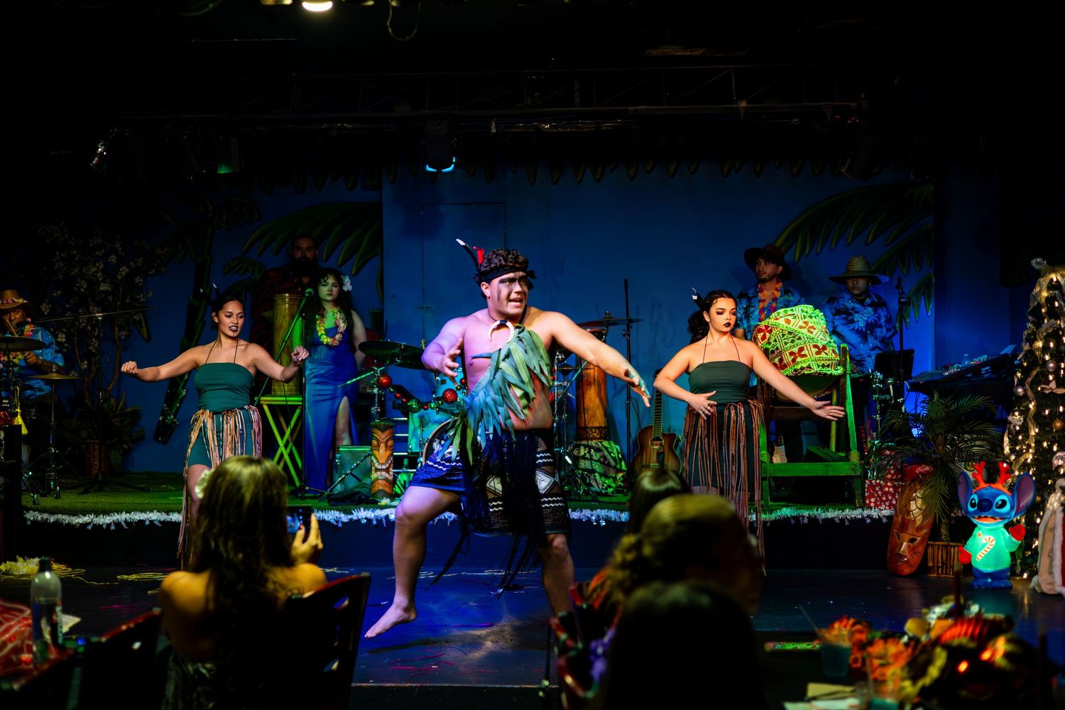 Performers in colorful costumes dance and act on a stage with vibrant lighting and theatrical scenery during a show.