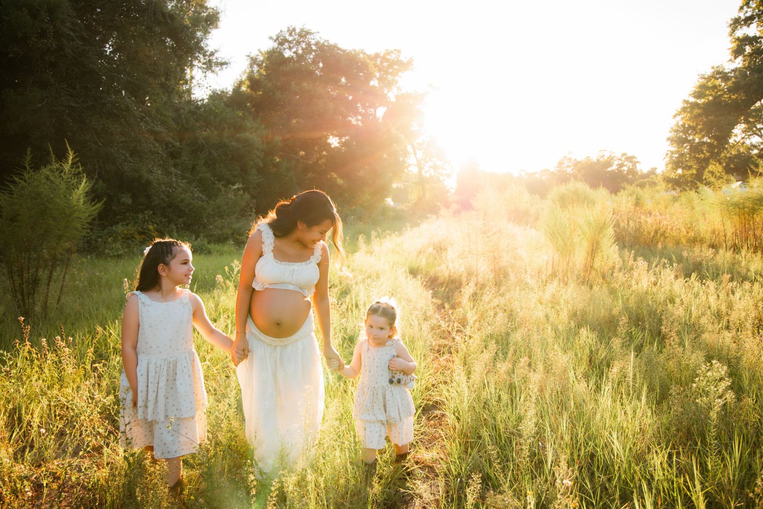 Pregnant woman in white dress walks with two daughters through sunlit green meadow during golden hour.